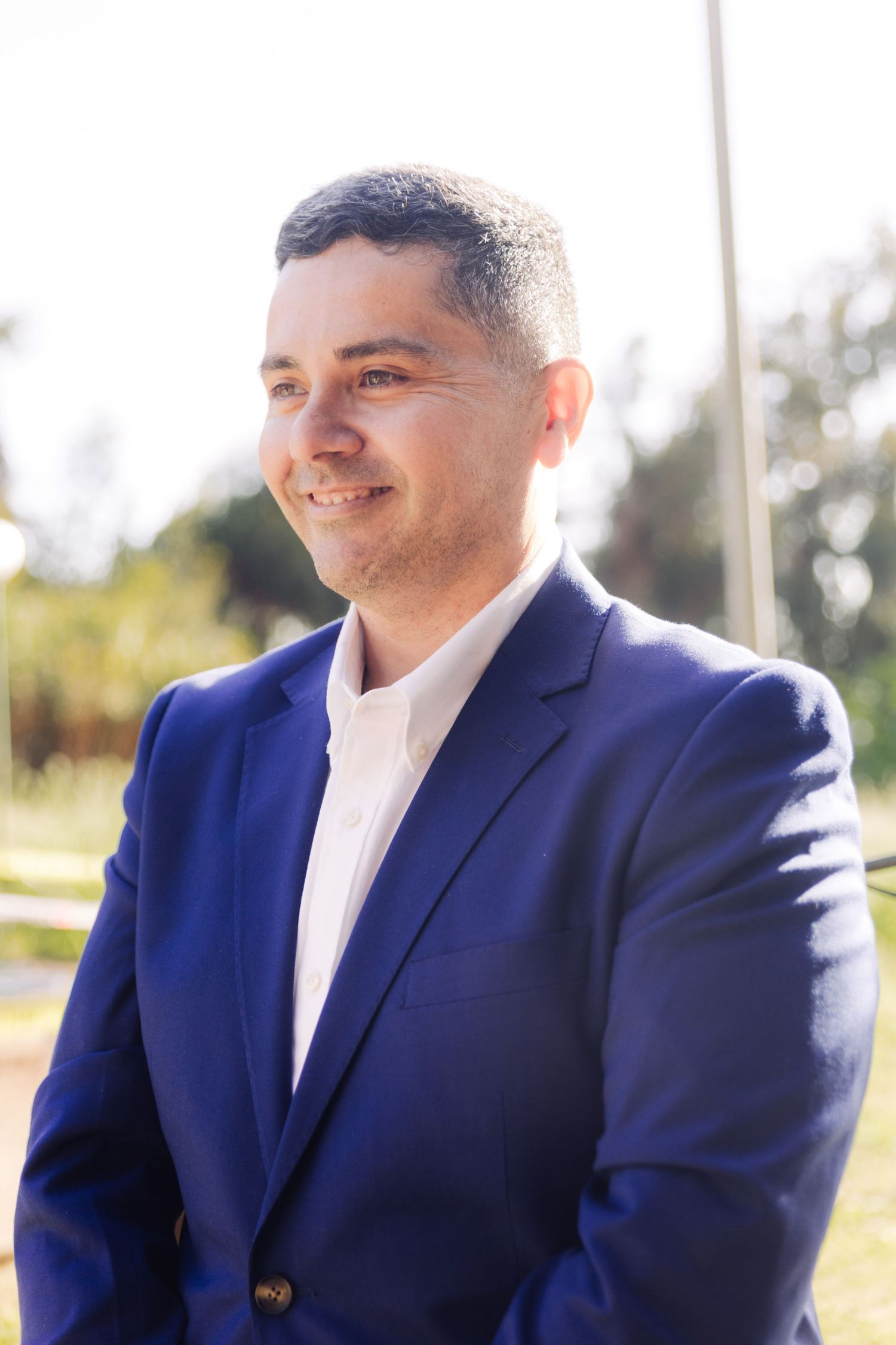 Smiling man in blue suit jacket and white shirt outdoors in bright sunlight.