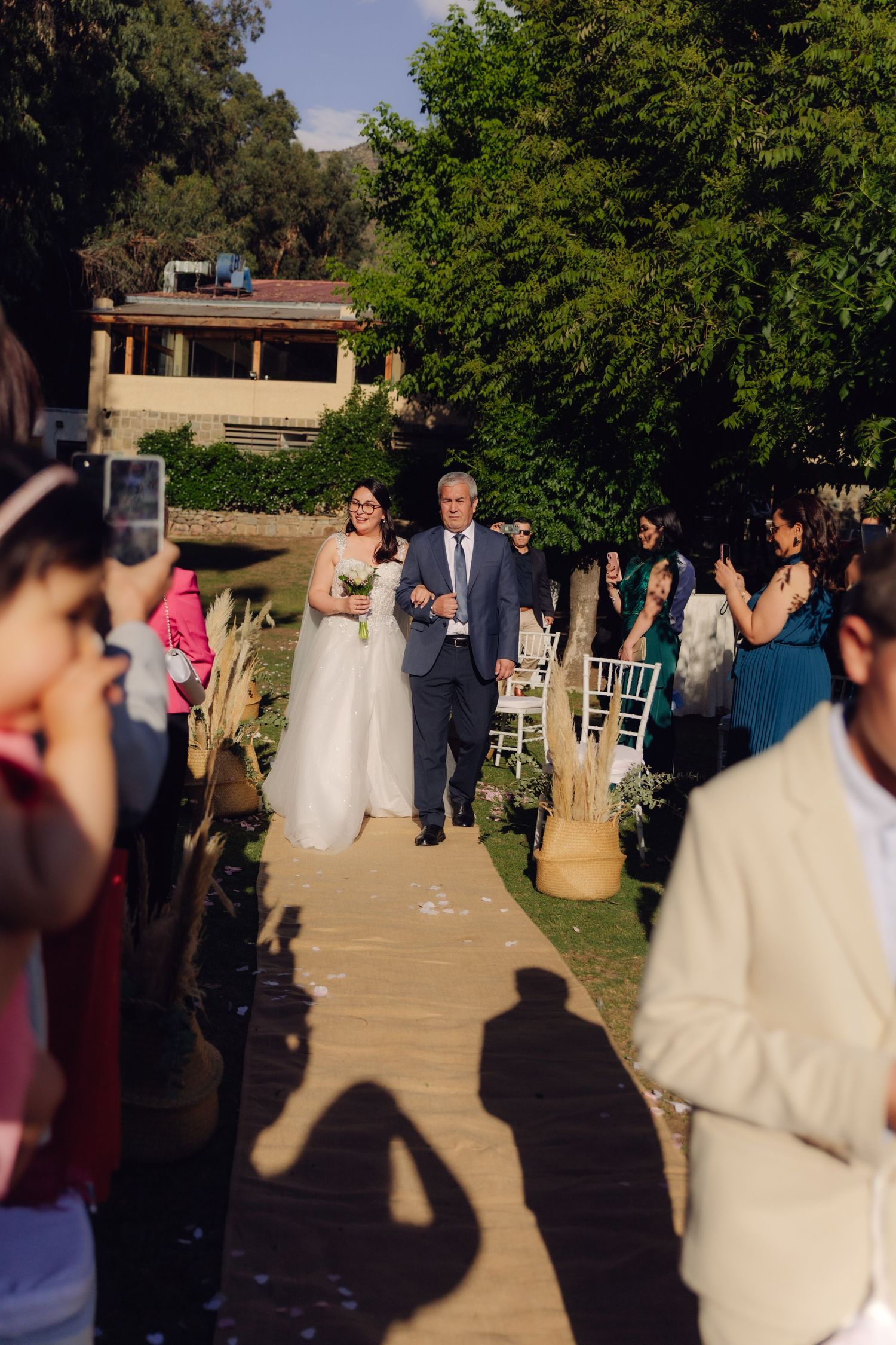 Bride walks down outdoor aisle with escort as guests applaud at garden wedding ceremony.