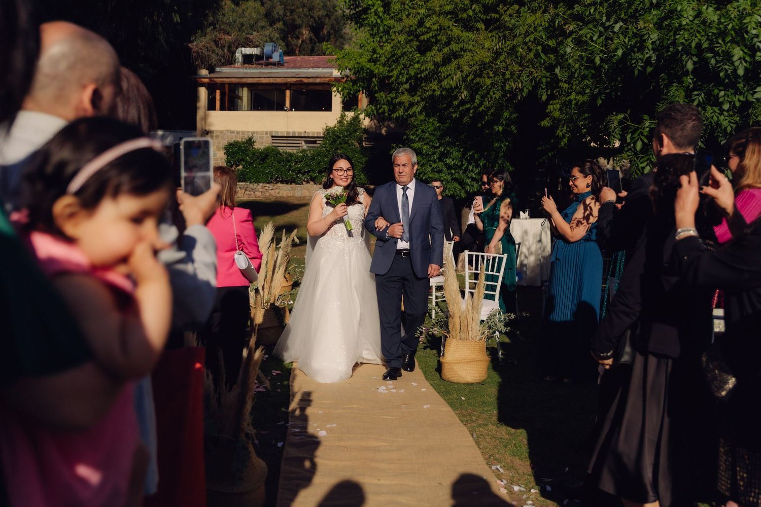 Bride and escort walk down sunlit outdoor aisle surrounded by applauding wedding guests.