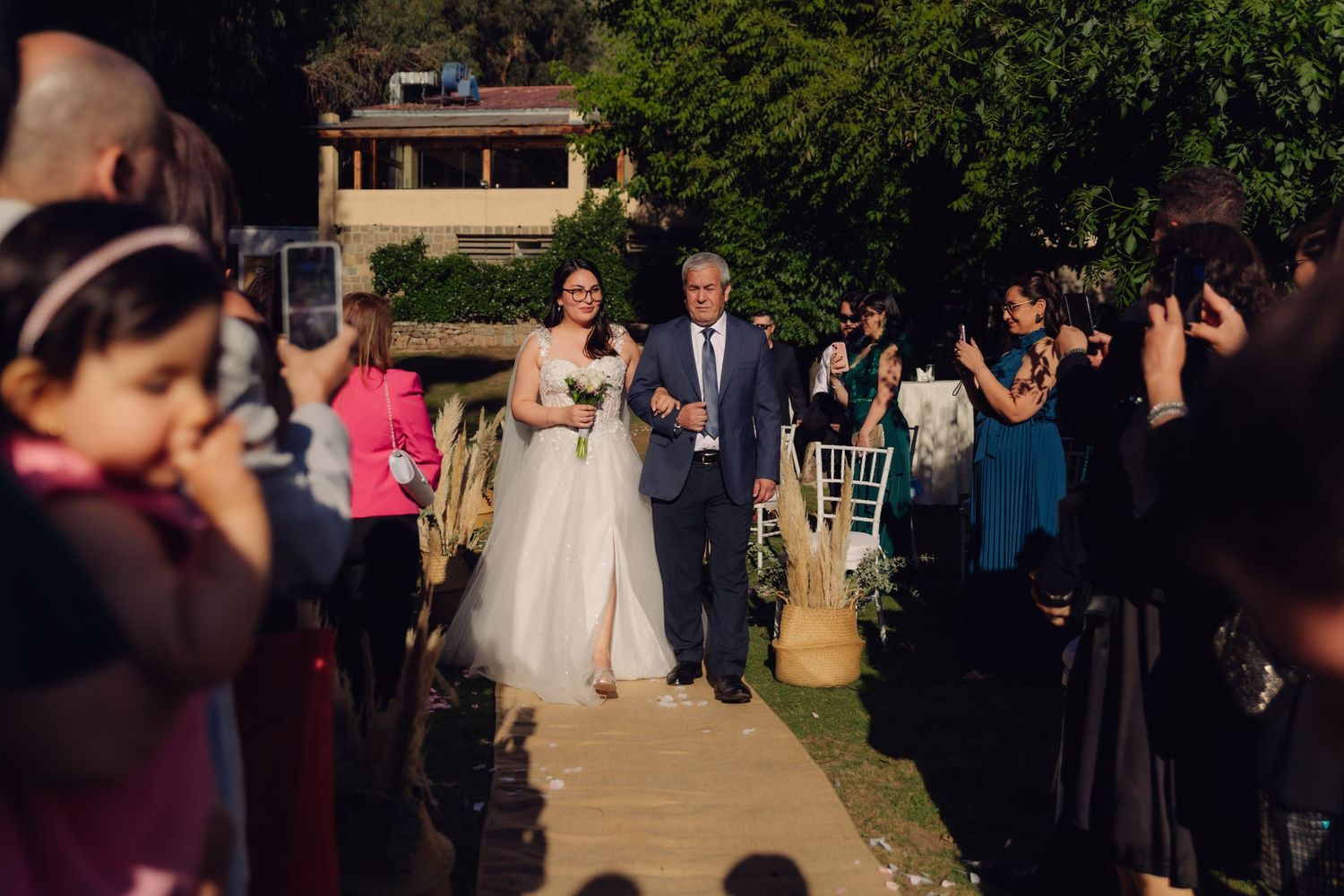 Bride in white gown and escort proceed down outdoor wedding aisle with cheering guests.