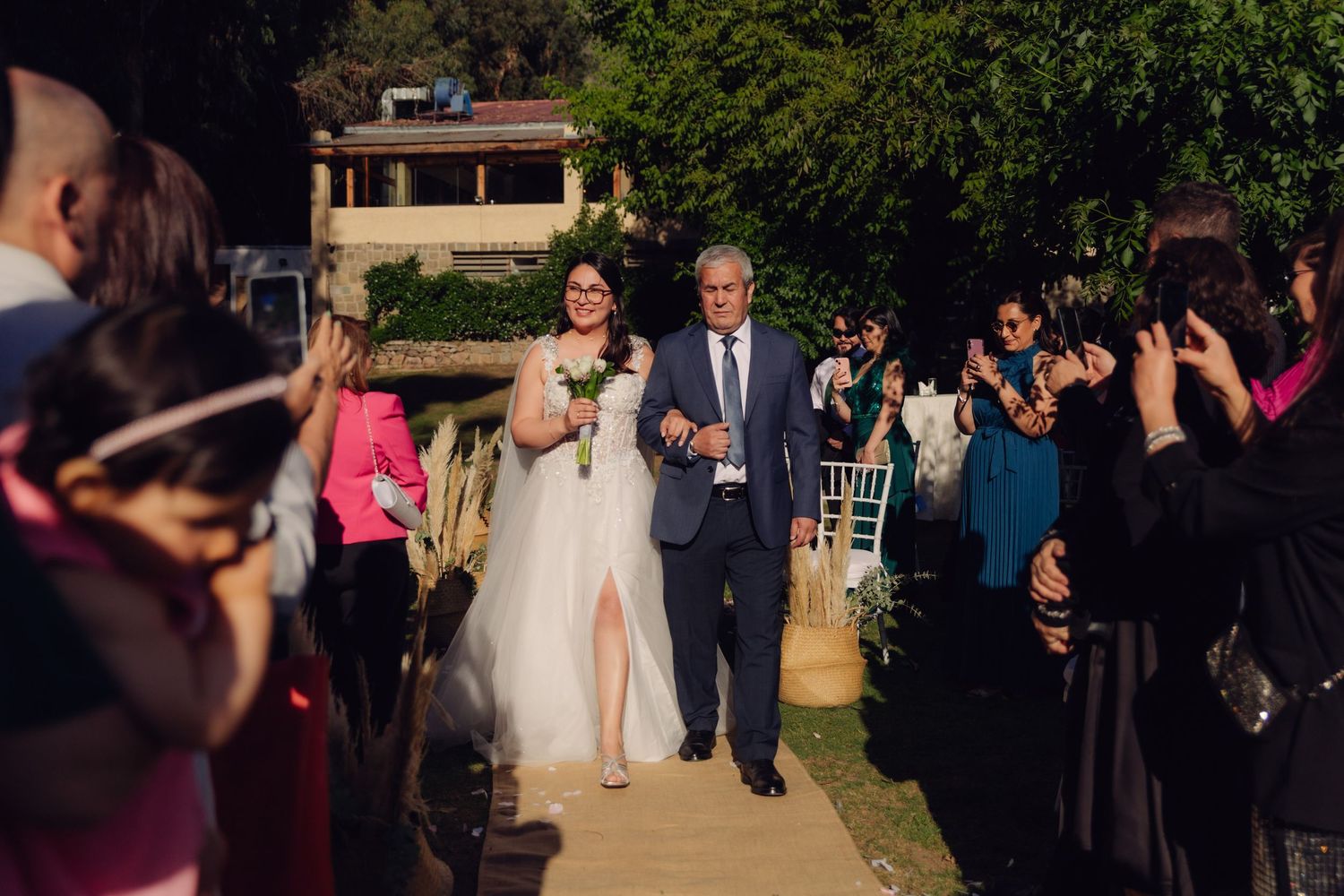 Bride and escort walk down garden wedding aisle as guests celebrate on both sides.
