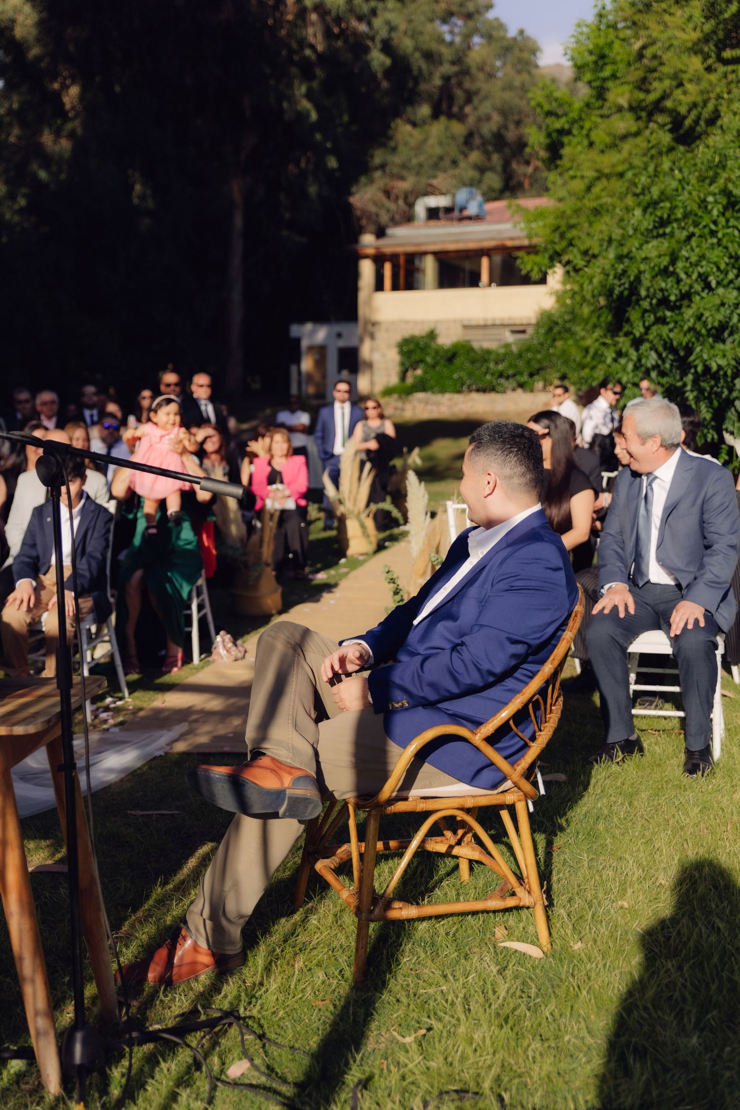 Wedding guest seated in rattan chair watches outdoor ceremony proceedings.
