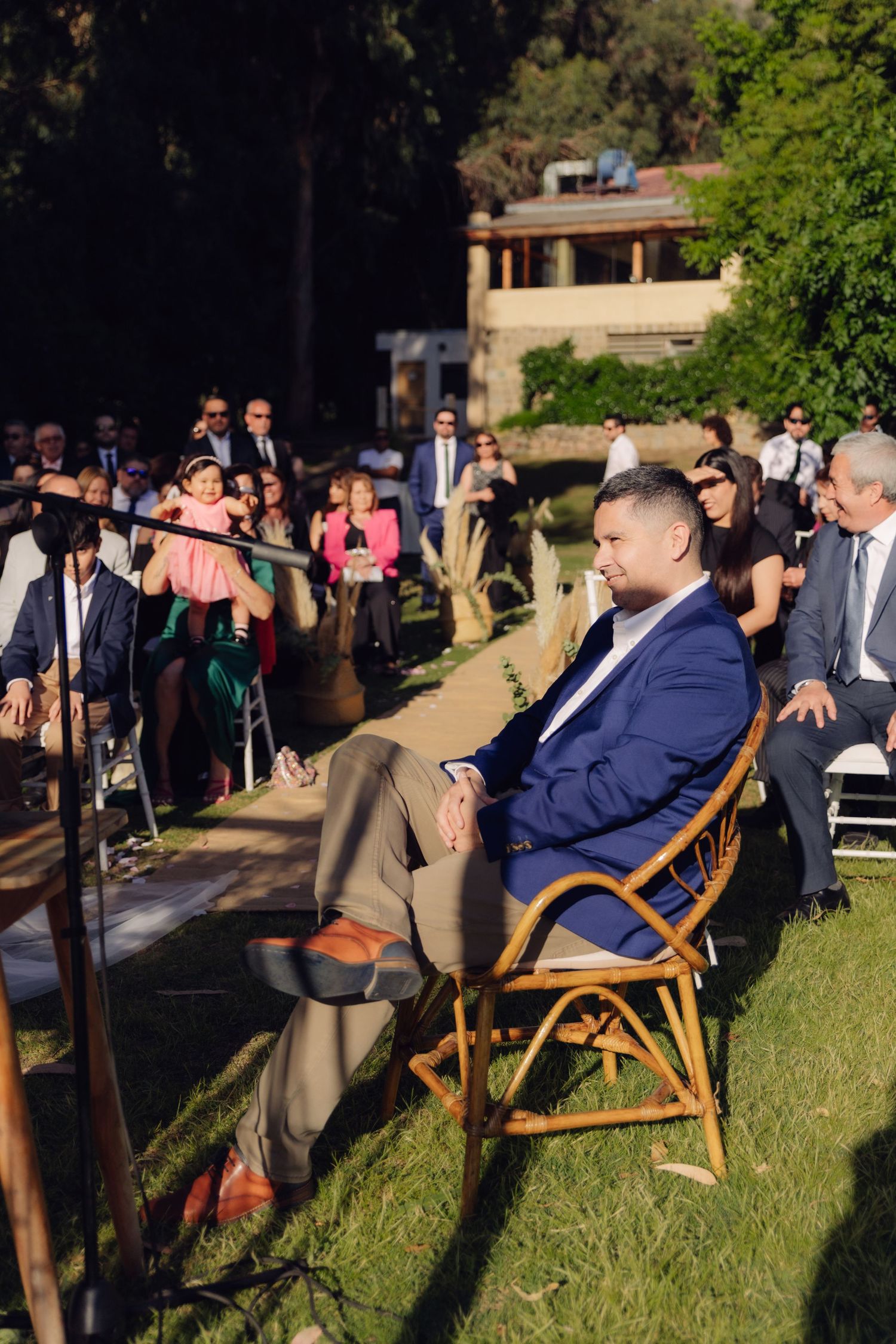 Smiling man in blue blazer seated at outdoor wedding ceremony.