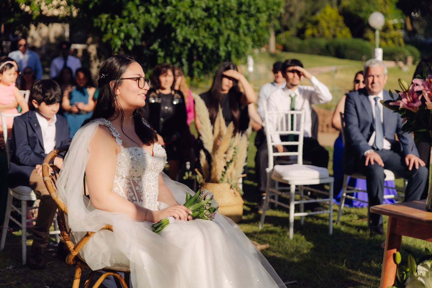 Bride in white gown smiles while seated at outdoor wedding.
