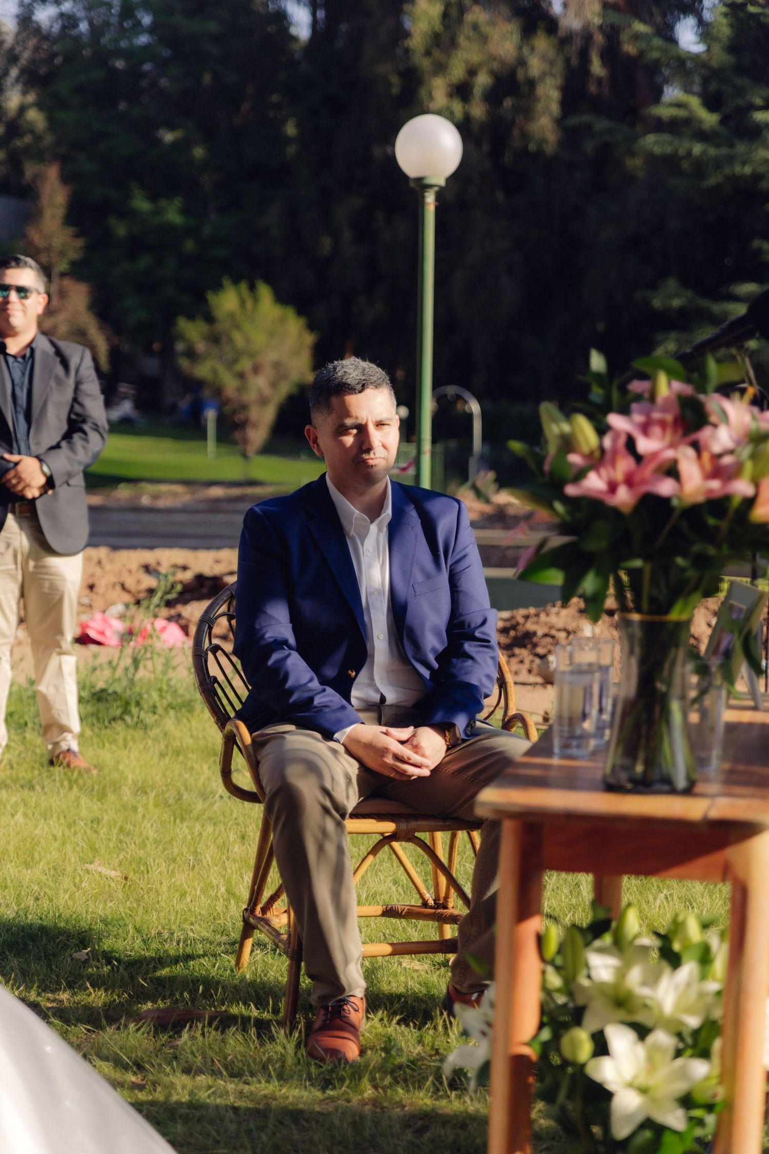 Groom in blue blazer sits pensively at outdoor garden ceremony.