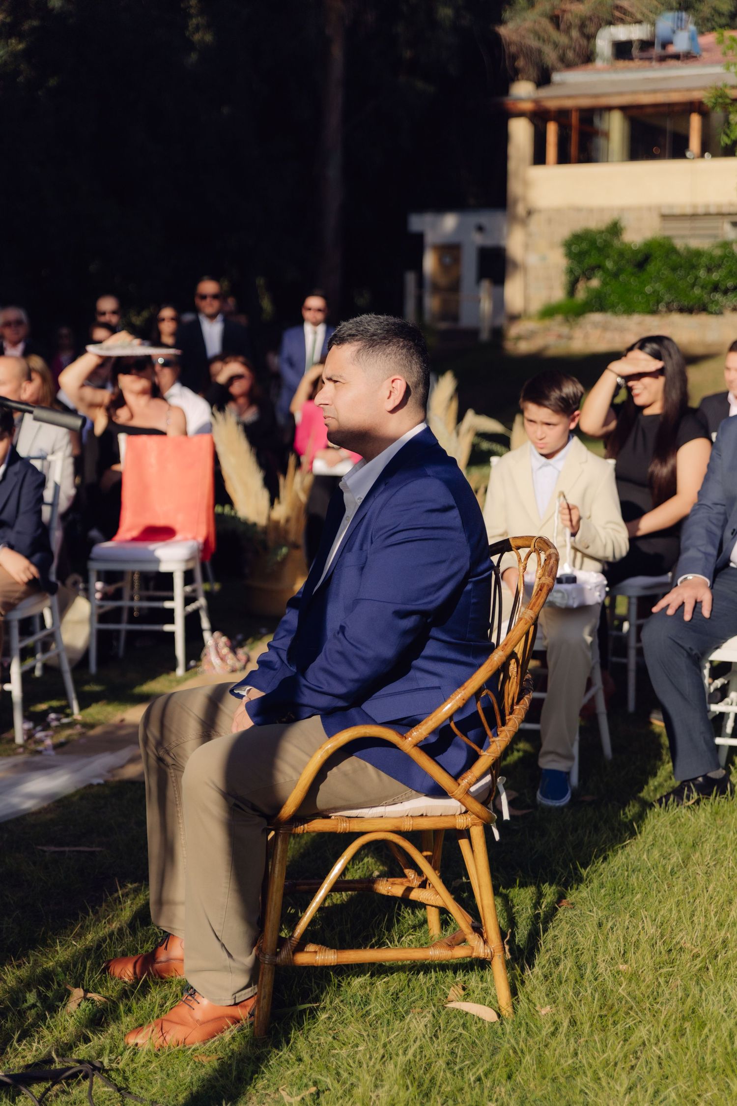 Wedding guest in navy blazer seated in rattan chair attentively watching outdoor ceremony with crowd behind.