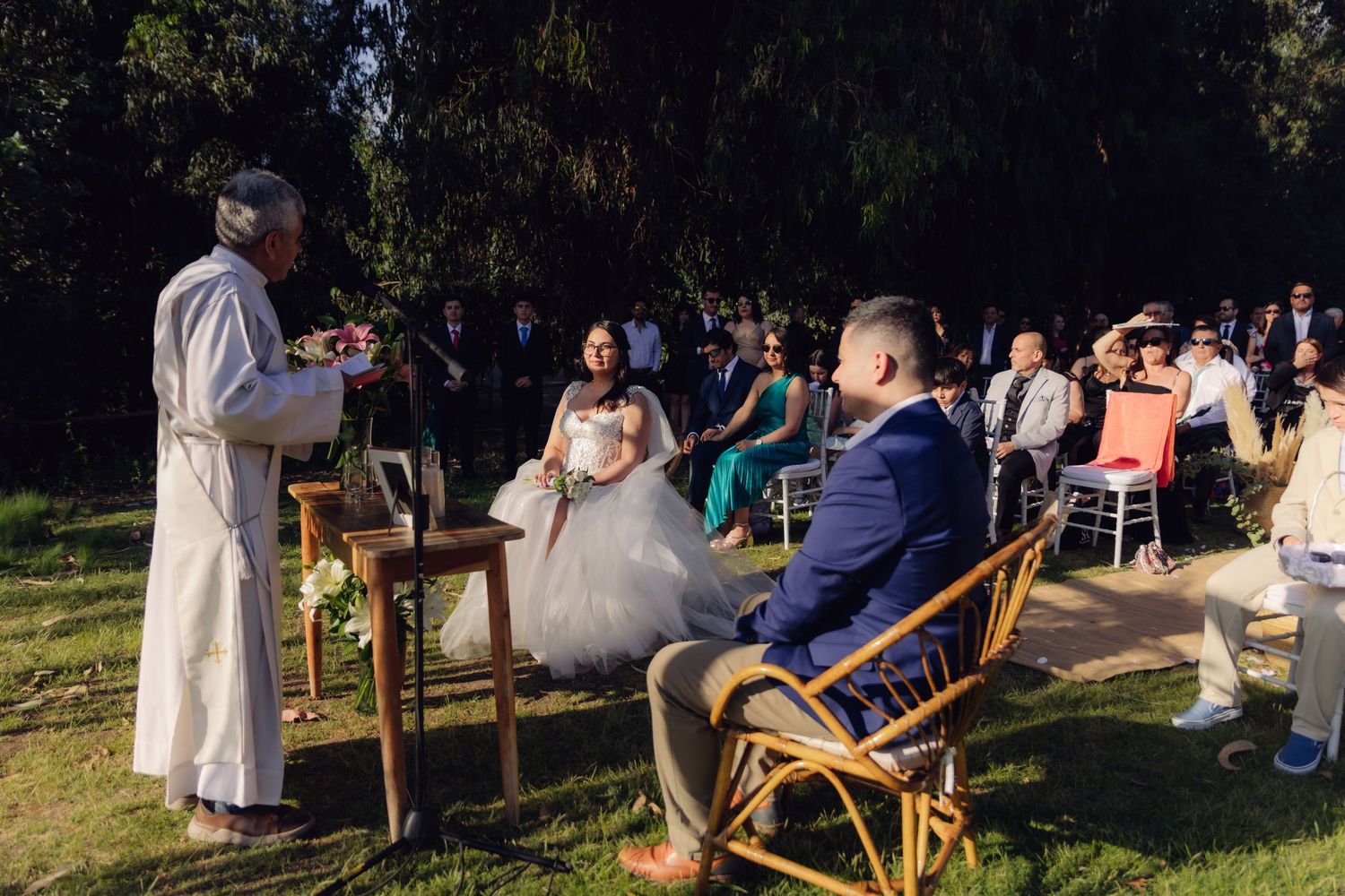 Outdoor wedding ceremony with bride in white gown and groom in blue suit seated before officiant at wooden altar.
