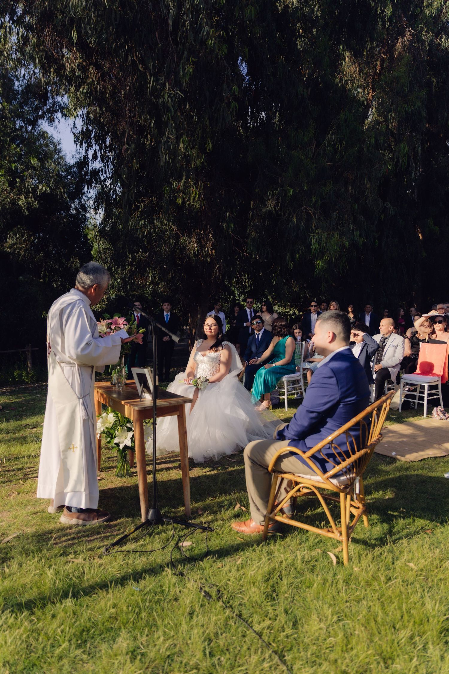 Wedding ceremony on green lawn with priest reading from book while bride and groom sit in rattan chairs.
