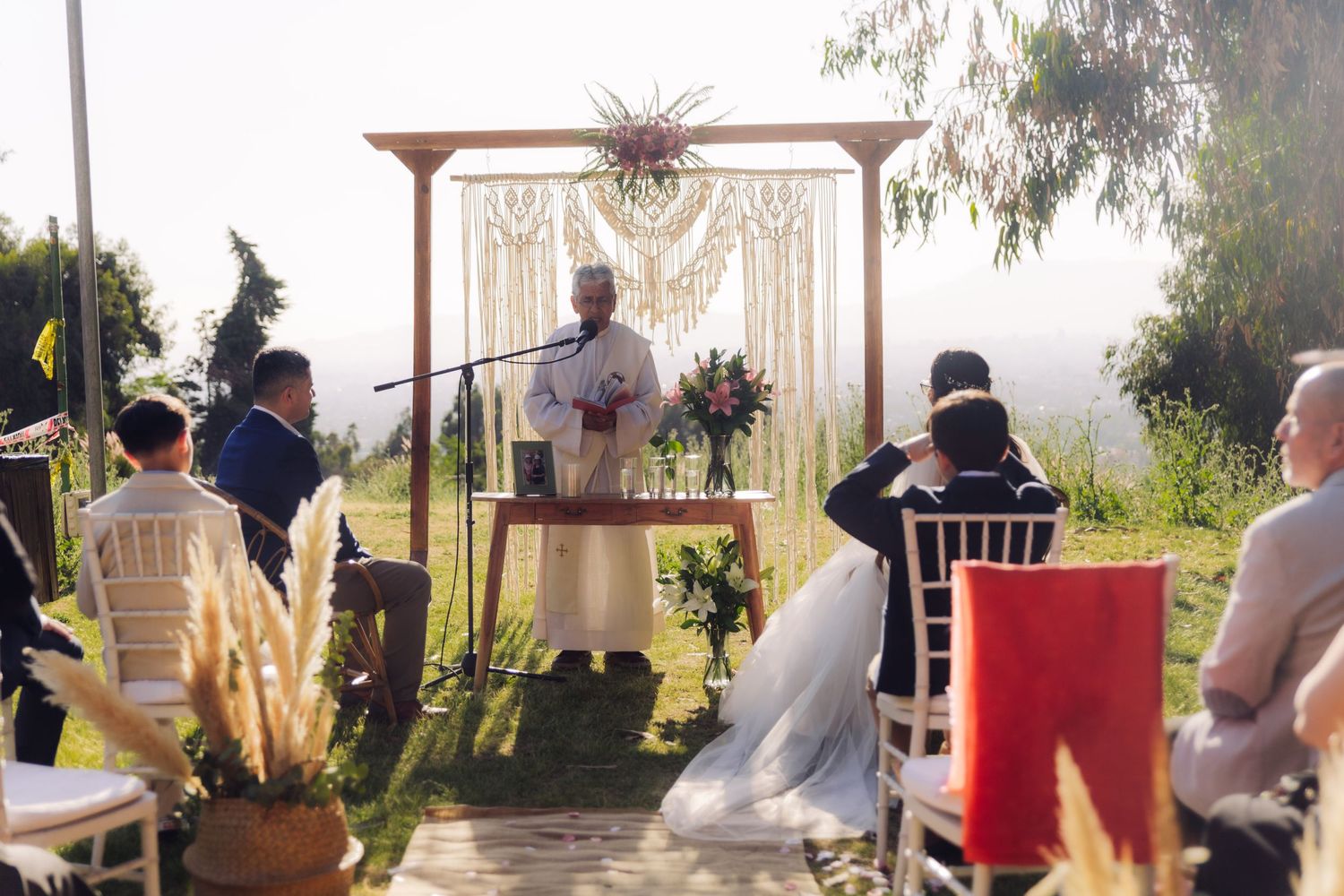 Outdoor wedding ceremony with officiant at boho macrame altar, guests seated on white chairs in sunlit garden.