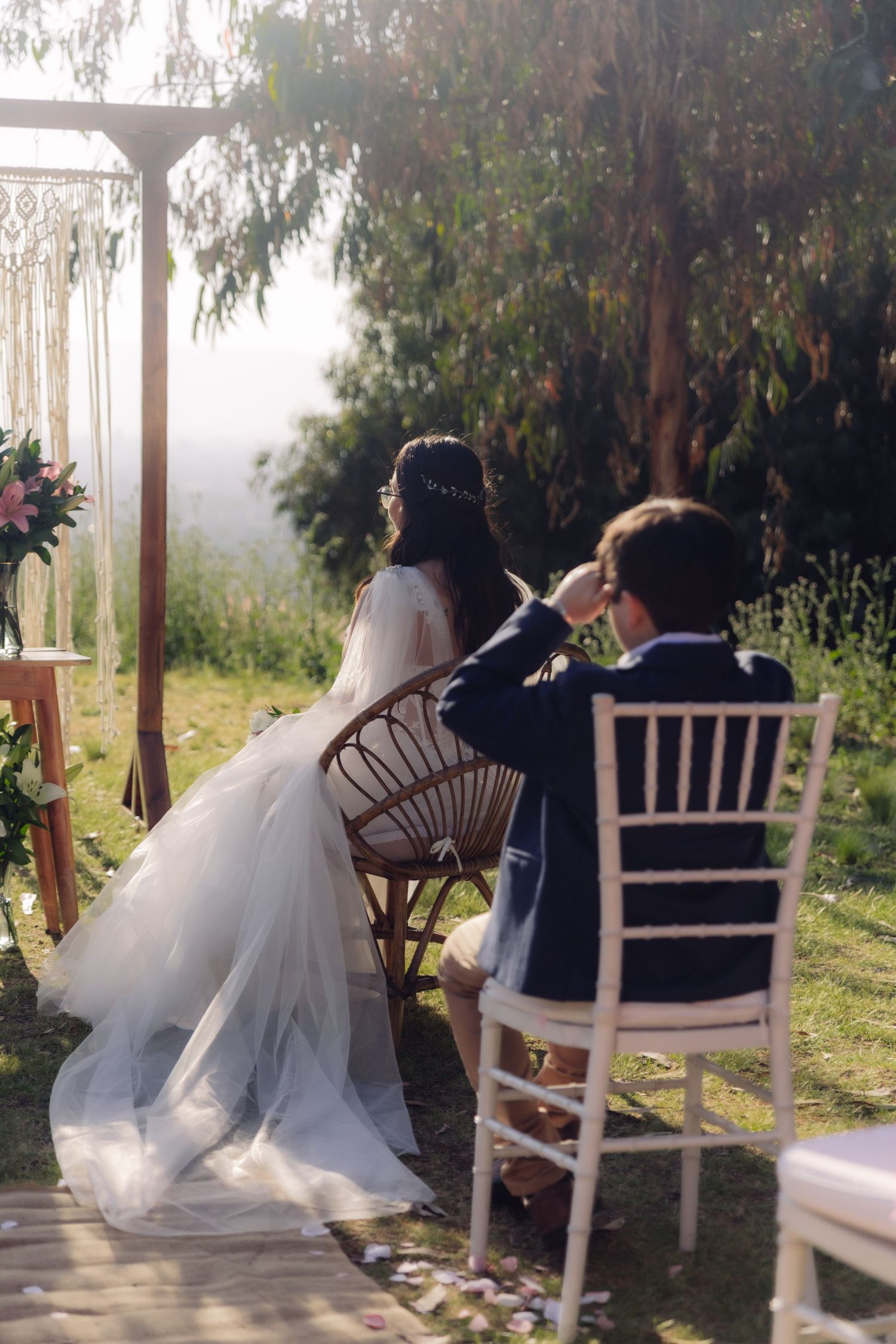Bride in flowing white gown and groom in navy suit seated at outdoor wedding ceremony near macrame arch.