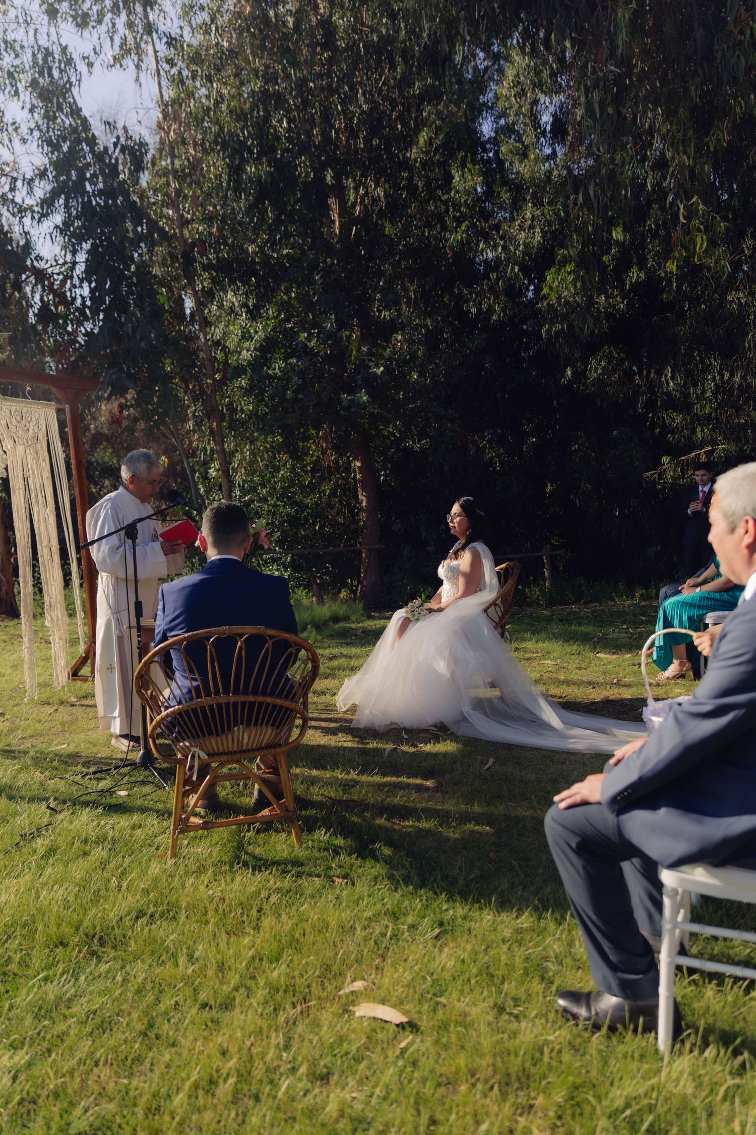 Outdoor wedding ceremony with bride in white gown smiling at officiant while groom and guests look on.