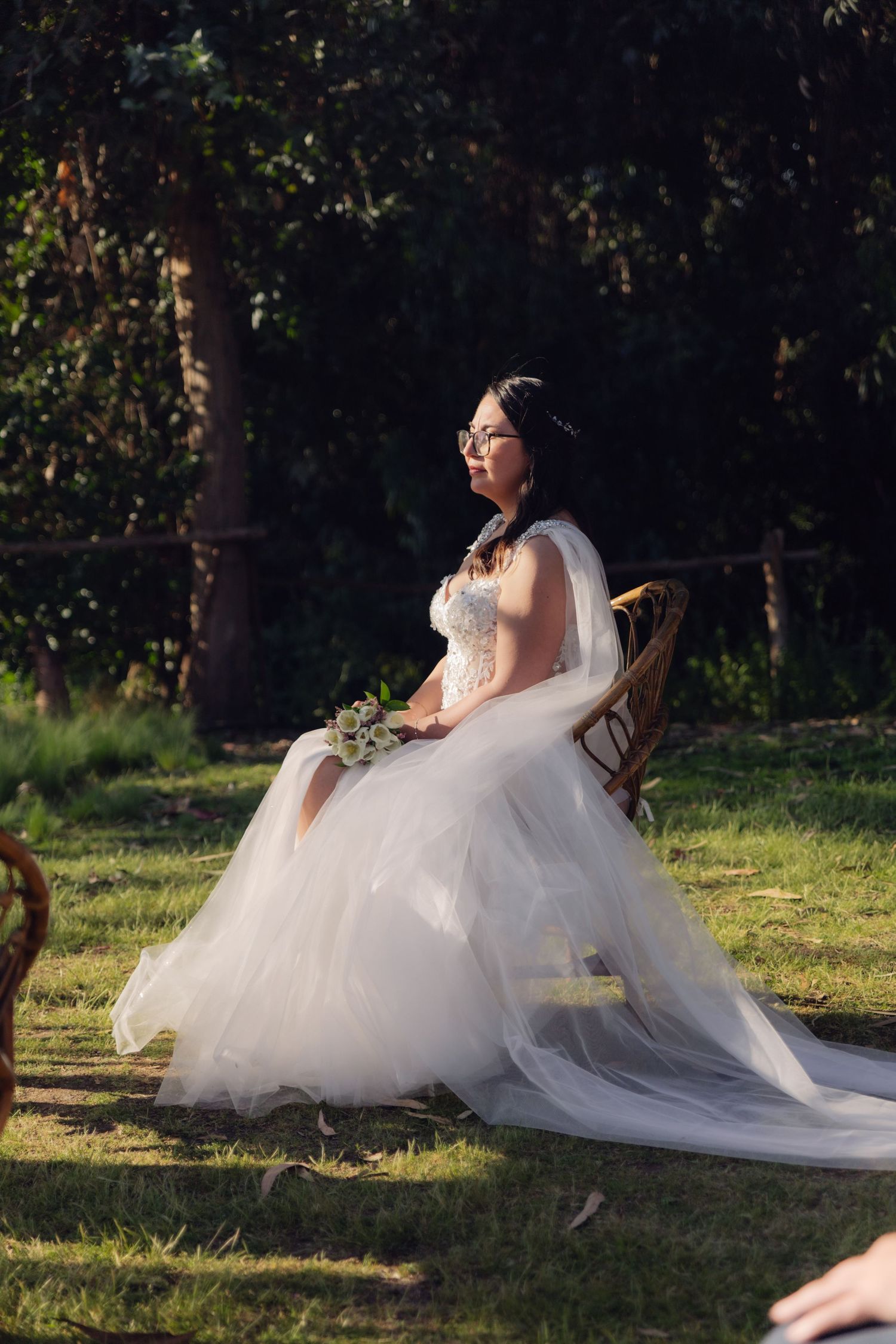 Elegant bride in white tulle ball gown with veil seated in rattan chair during outdoor garden ceremony.
