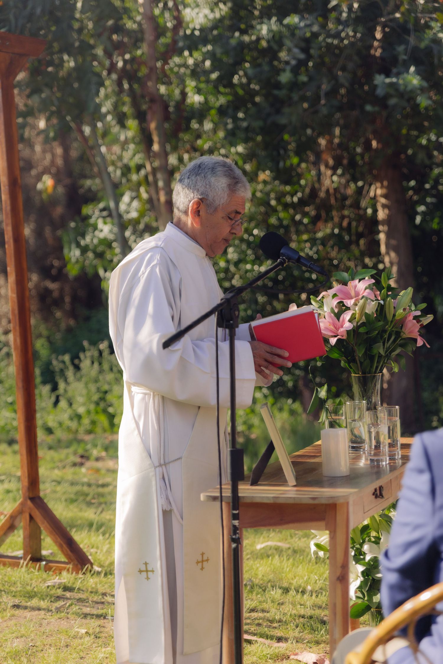 Priest in white vestments reads from red book at wooden altar adorned with pink lilies outdoors.