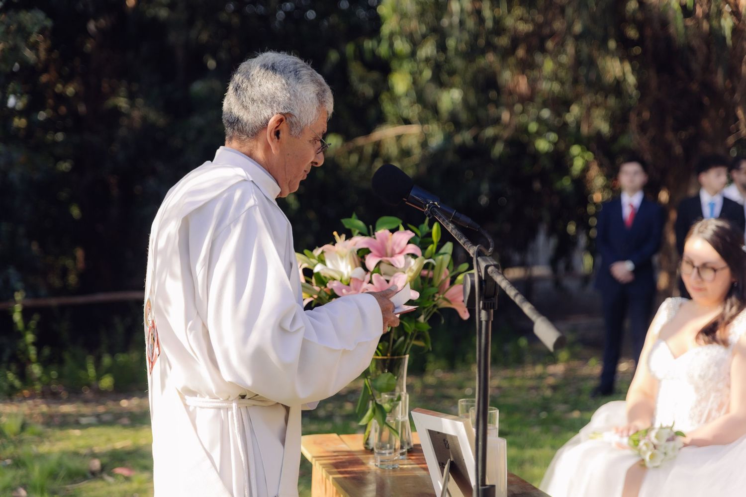 Officiant in white robes holds pink lily bouquet at microphone while bride in white gown watches ceremony.