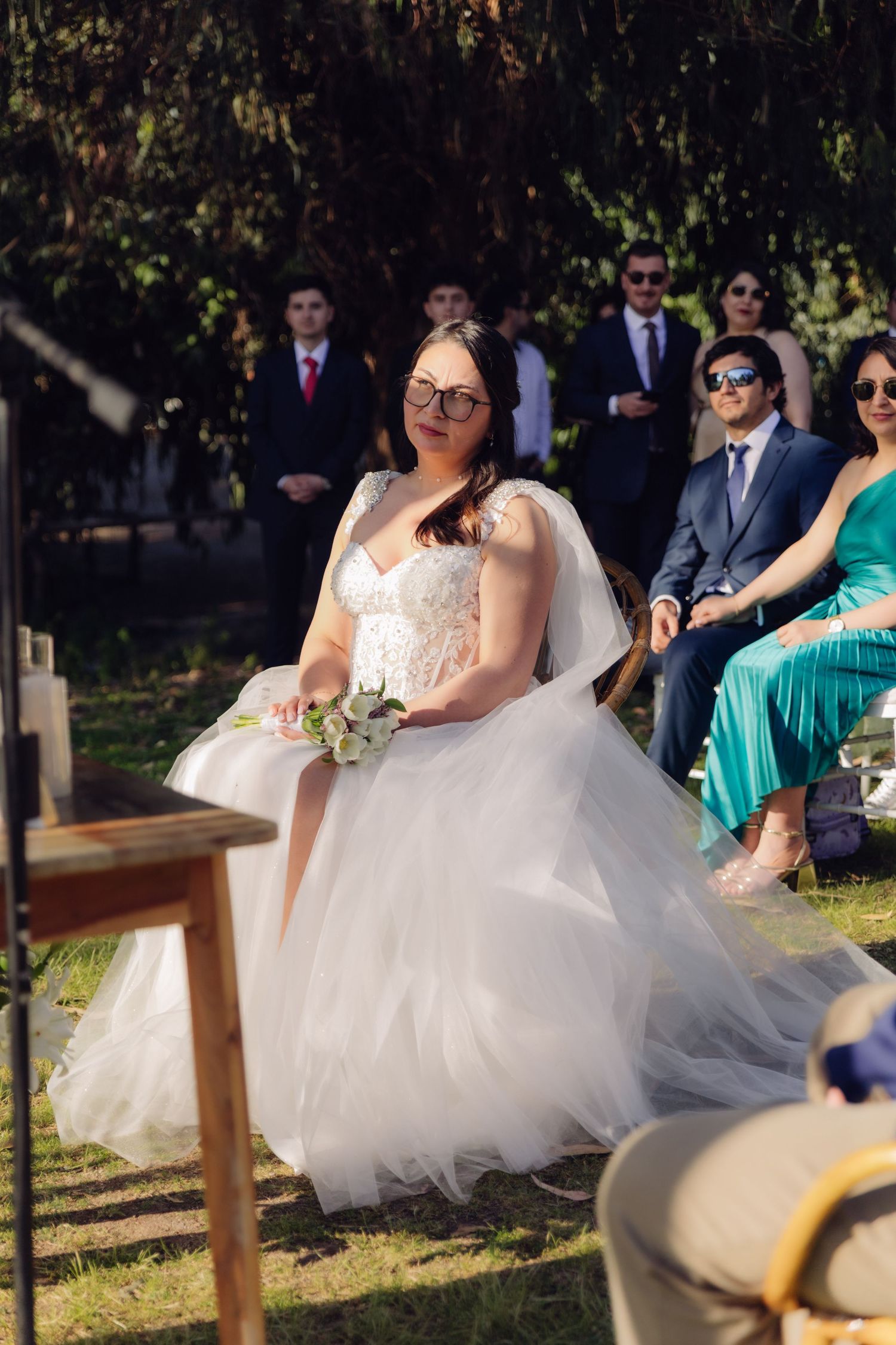 Smiling bride in white tulle gown with glasses holds bouquet during outdoor wedding ceremony with guests.