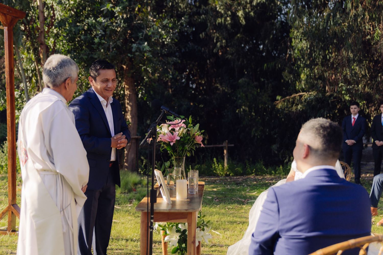 Outdoor wedding ceremony with officiant and groom at altar as guests watch in a garden setting.