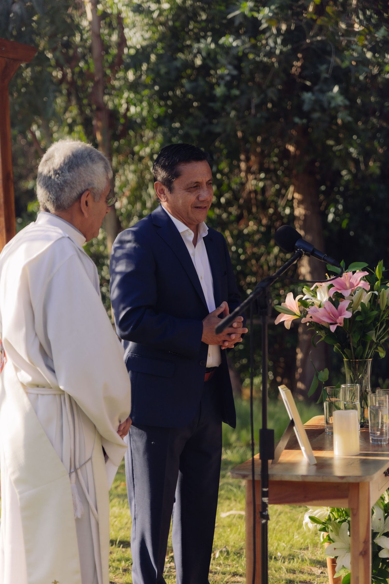Man in navy suit speaking at outdoor wedding ceremony with officiant in white robe and pink lilies nearby.