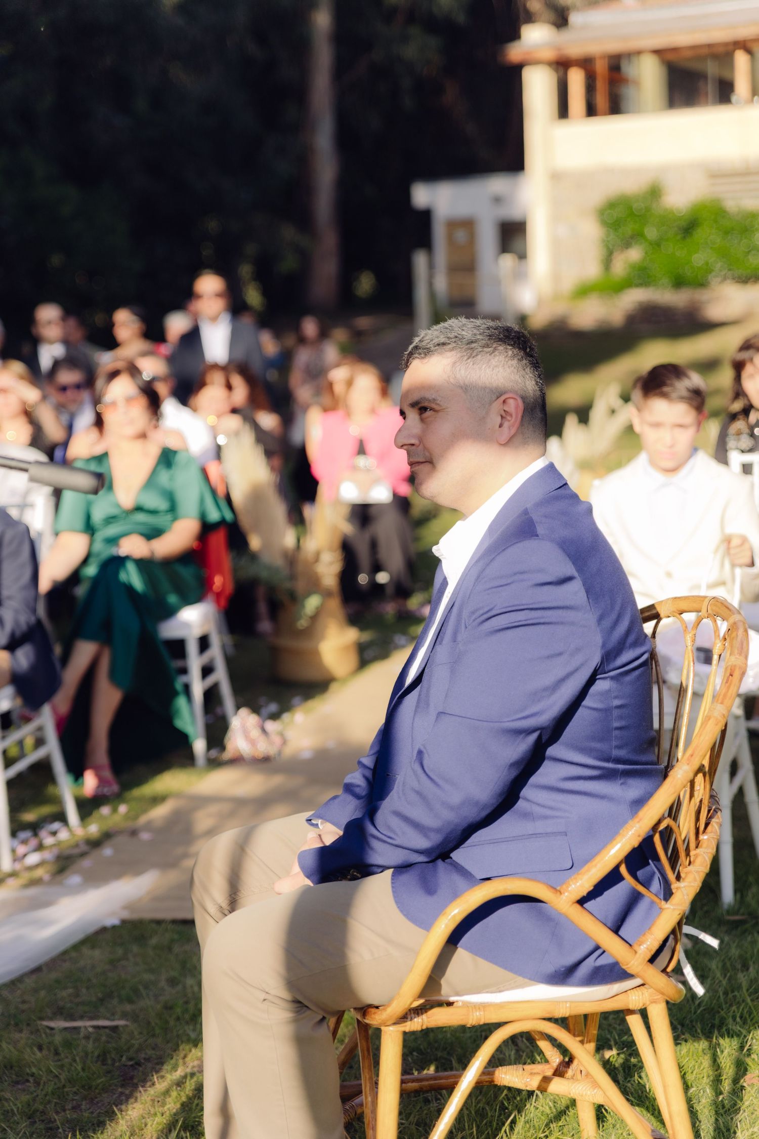 Man in blue suit seated in rattan chair watching outdoor wedding ceremony with guests behind him.