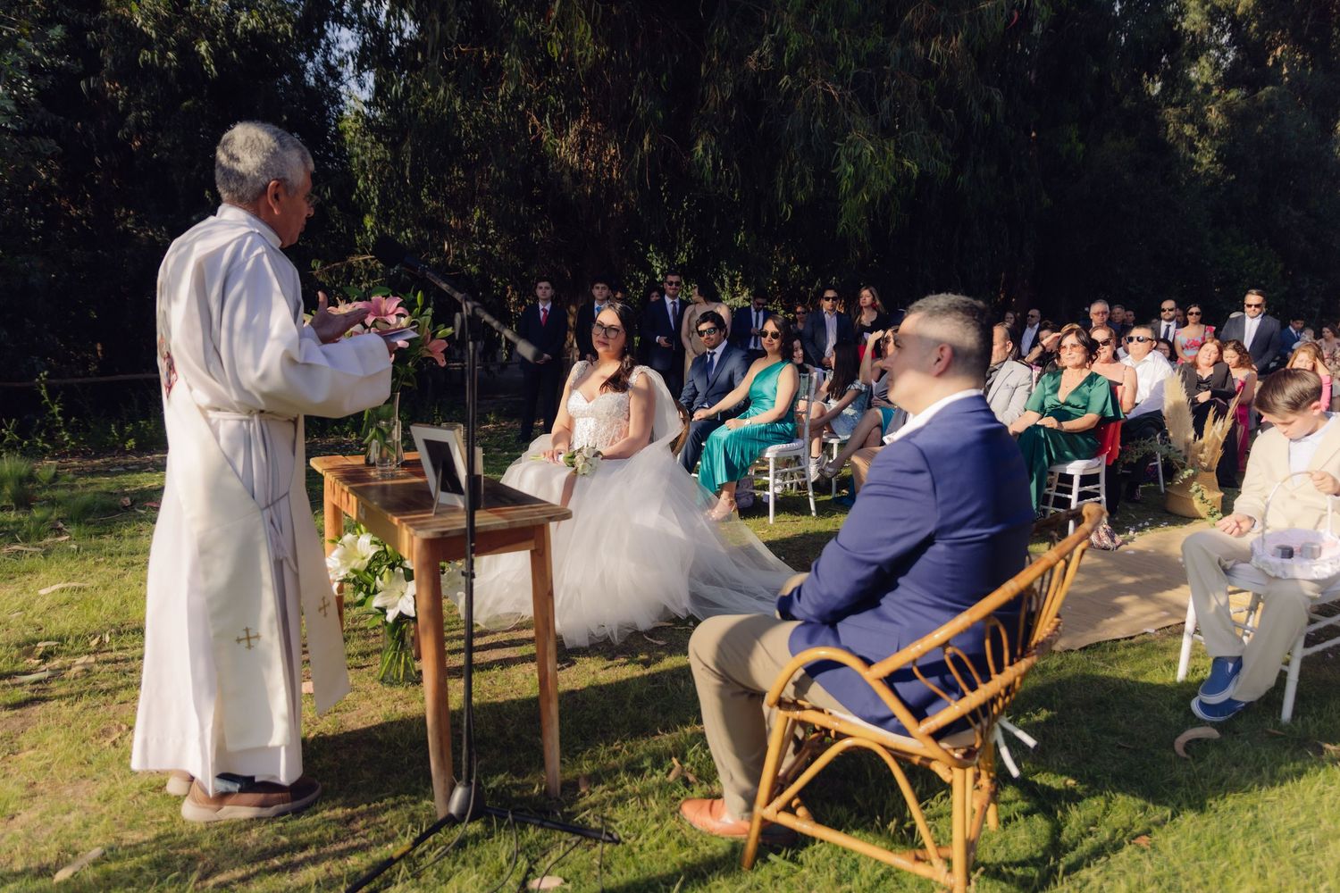 Officiant in white robe leads outdoor wedding ceremony with bride and groom seated before guests.