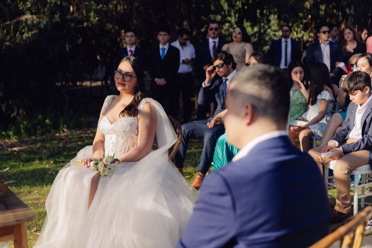 Bride in white gown with bouquet seated at outdoor wedding ceremony surrounded by guests.