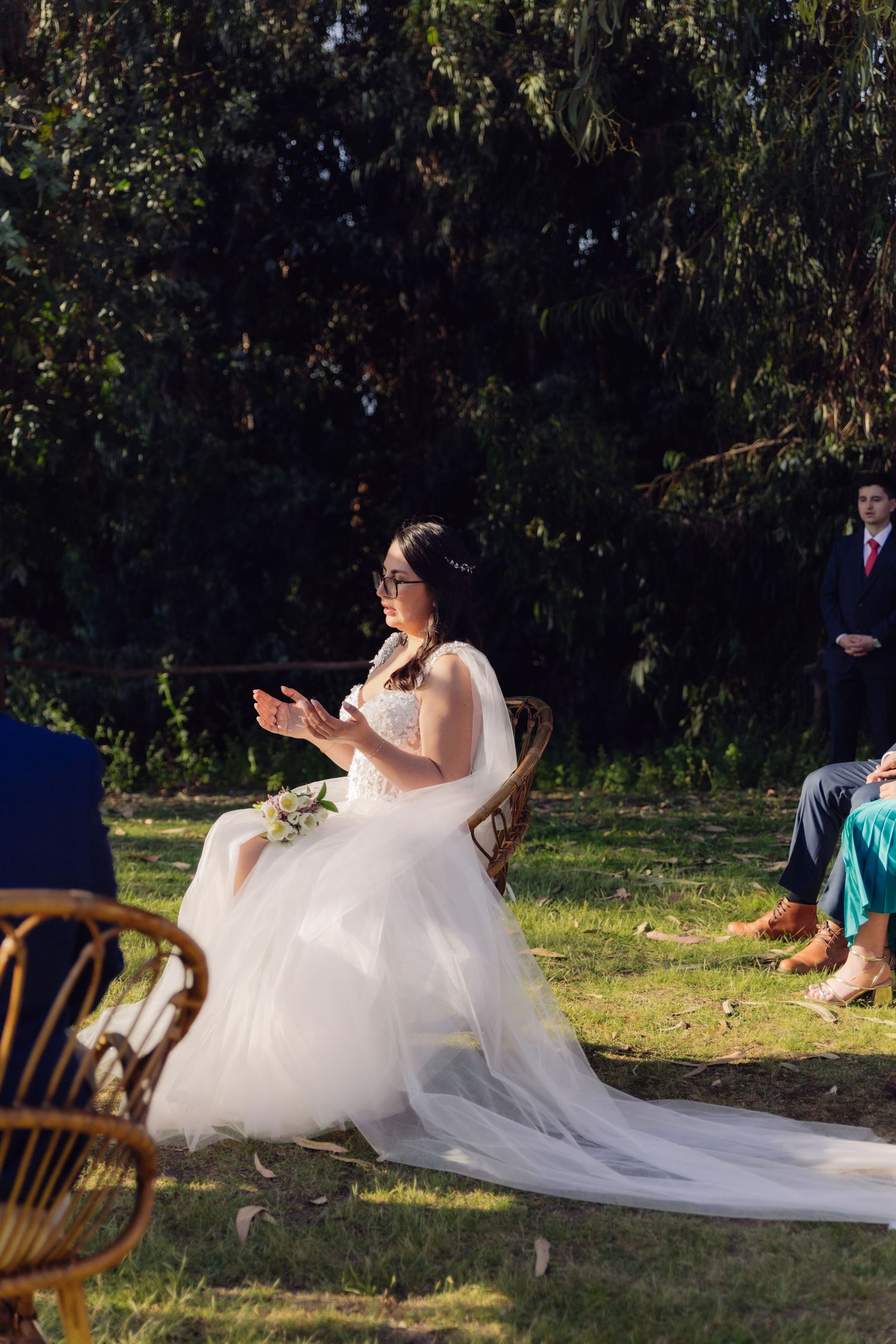Bride in elegant white tulle gown seated outdoors during wedding ceremony in a sunlit garden.