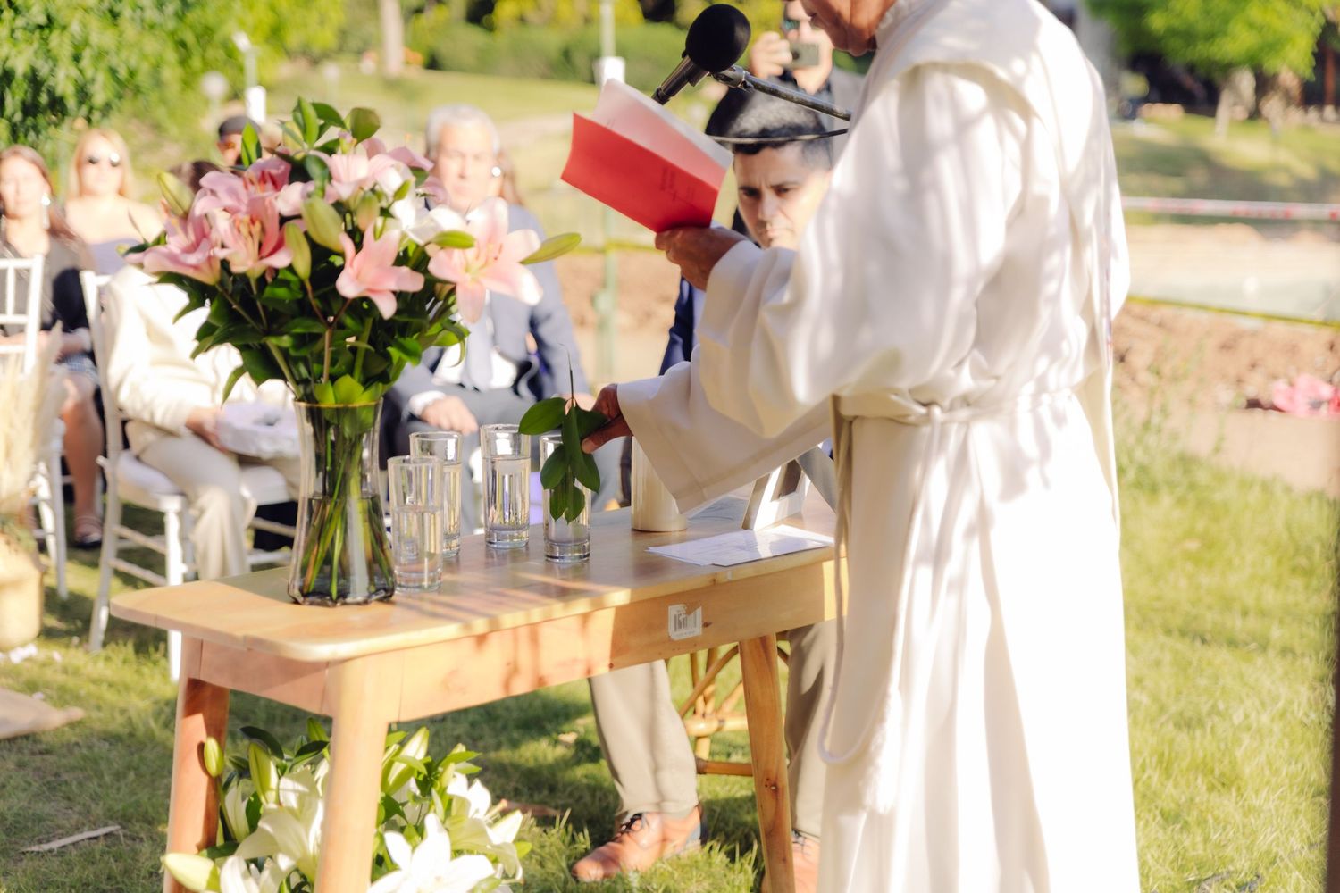 Officiant in white robe reads from red book at outdoor wedding ceremony altar with floral arrangements.
