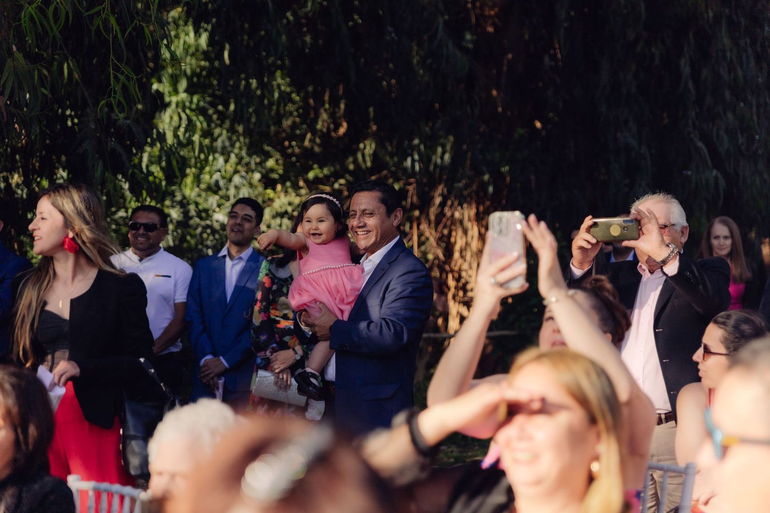 Wedding guests smile and take photos on smartphones during an outdoor garden ceremony.