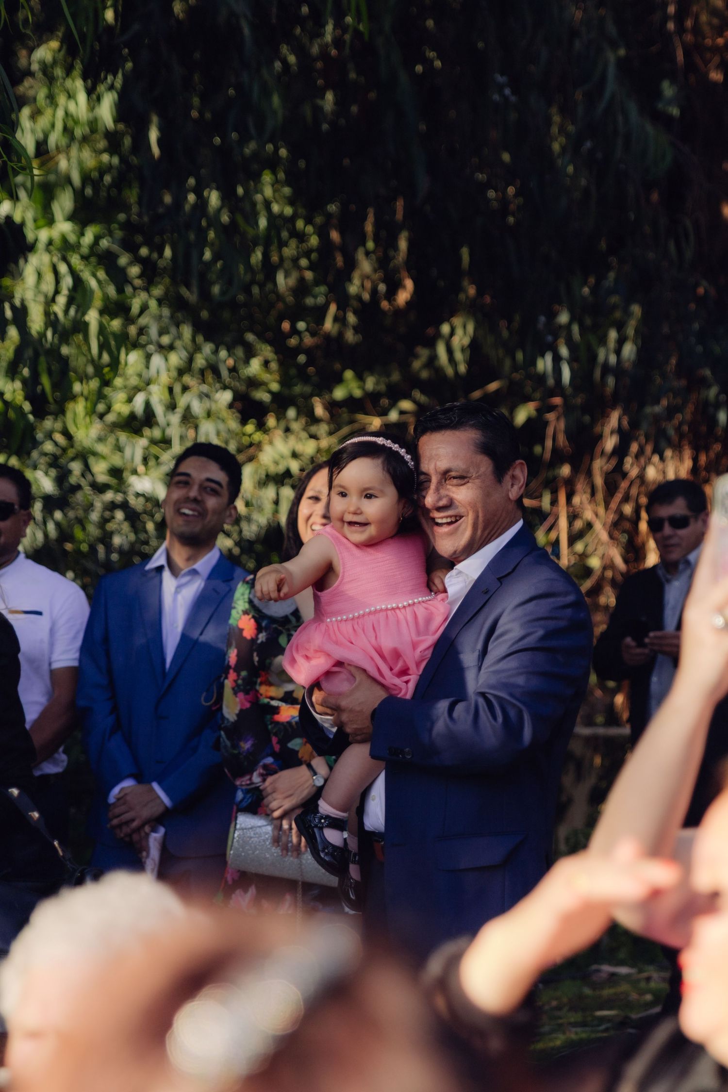 Smiling man in blue suit holds happy baby girl in pink dress at outdoor wedding celebration.