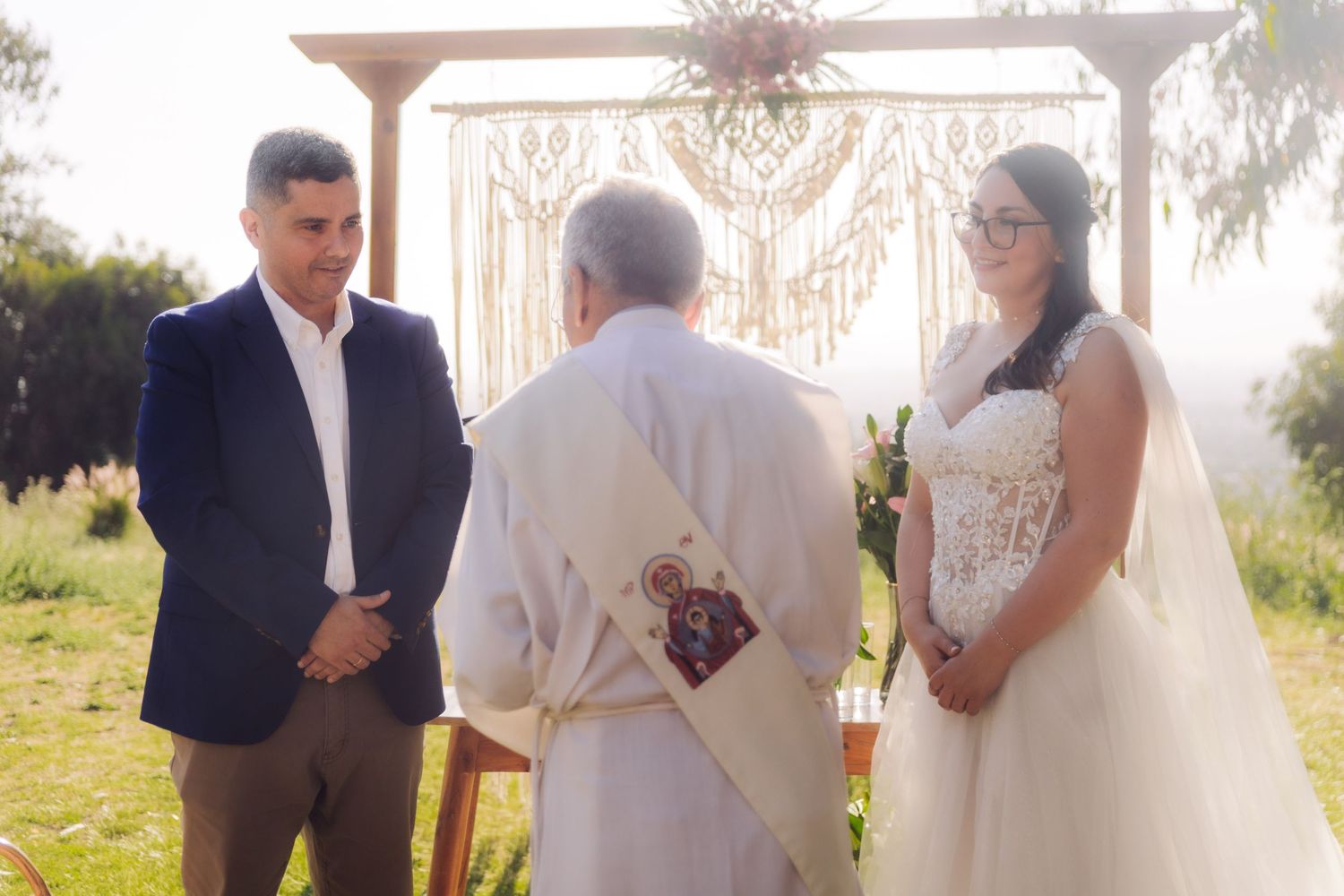 Bride in white gown and groom in blue suit exchange vows with officiant at outdoor wedding.