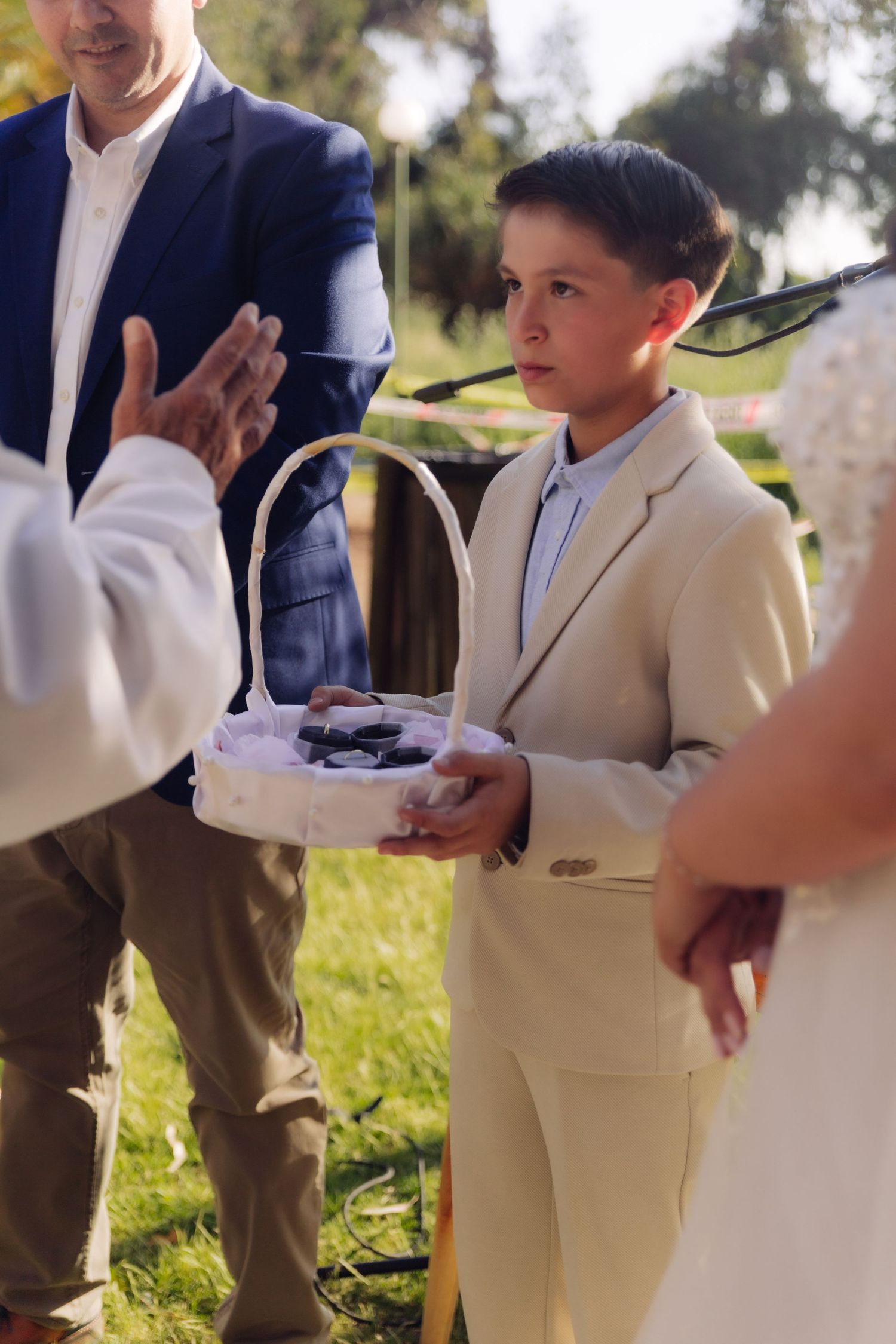 Young ring bearer in beige suit presents wedding rings in basket to officiant at outdoor ceremony.