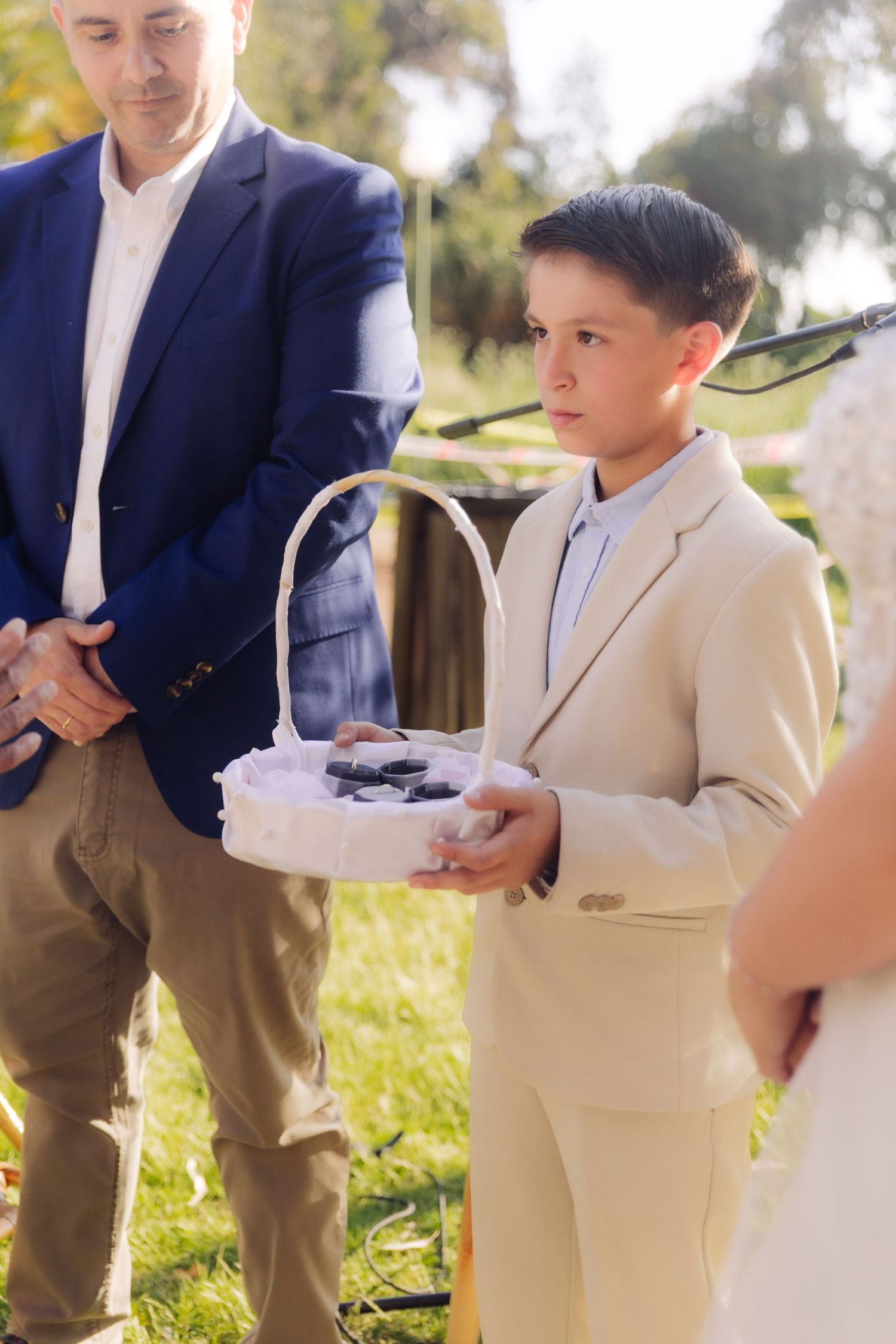 Serious young ring bearer in beige suit holds white basket with wedding rings at outdoor ceremony.