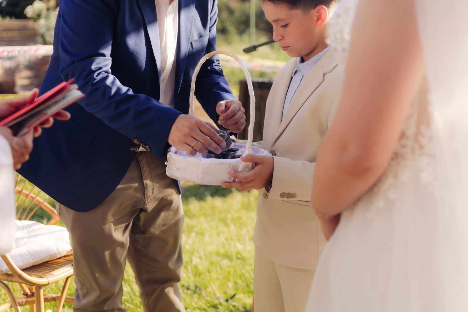 Groom in blue suit retrieves wedding rings from basket held by ring bearer at outdoor ceremony.
