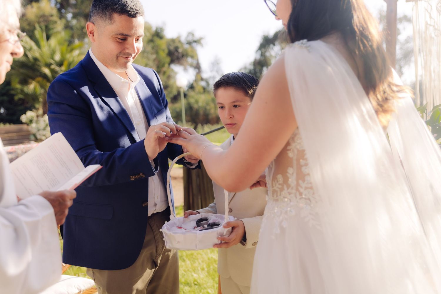 Smiling groom places wedding ring on brides finger assisted by ring bearer at outdoor ceremony.