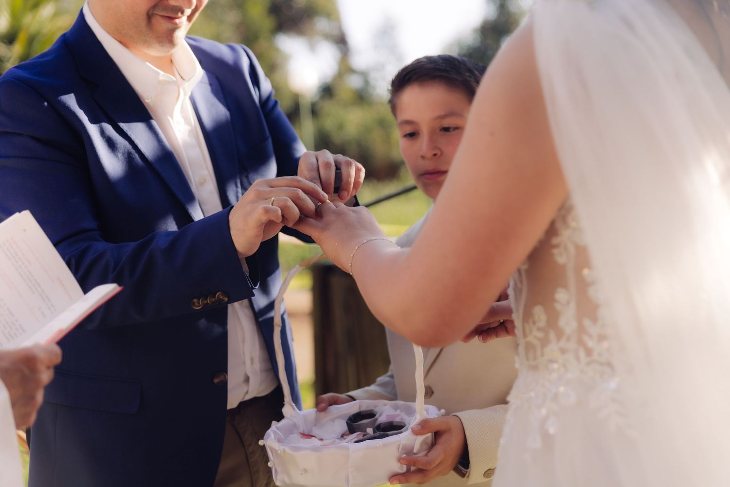 Groom places wedding ring on brides hand while ring bearer assists at sunny outdoor ceremony.