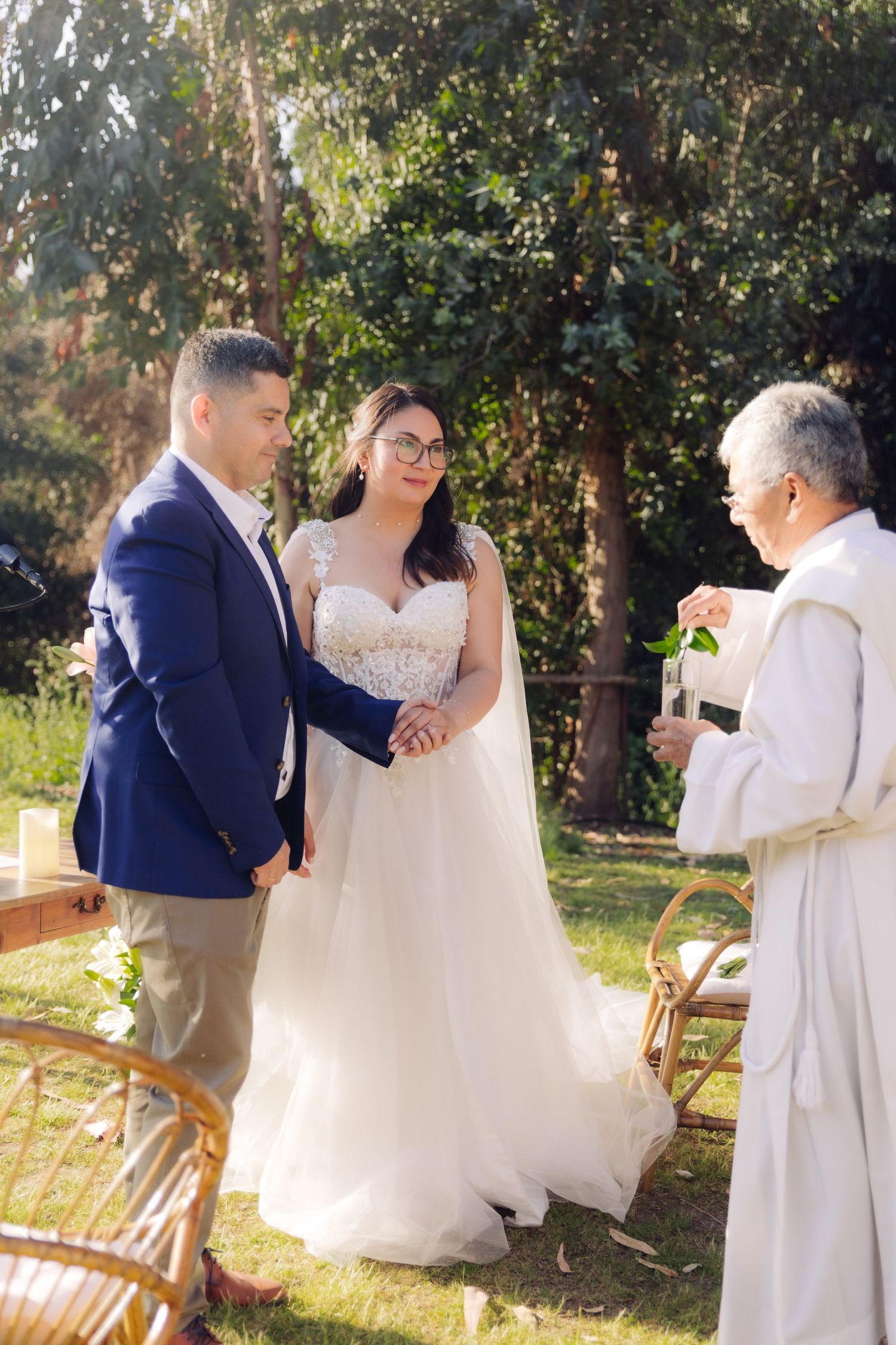 Bride and groom hold hands while elderly officiant in white robe blesses them at outdoor wedding.