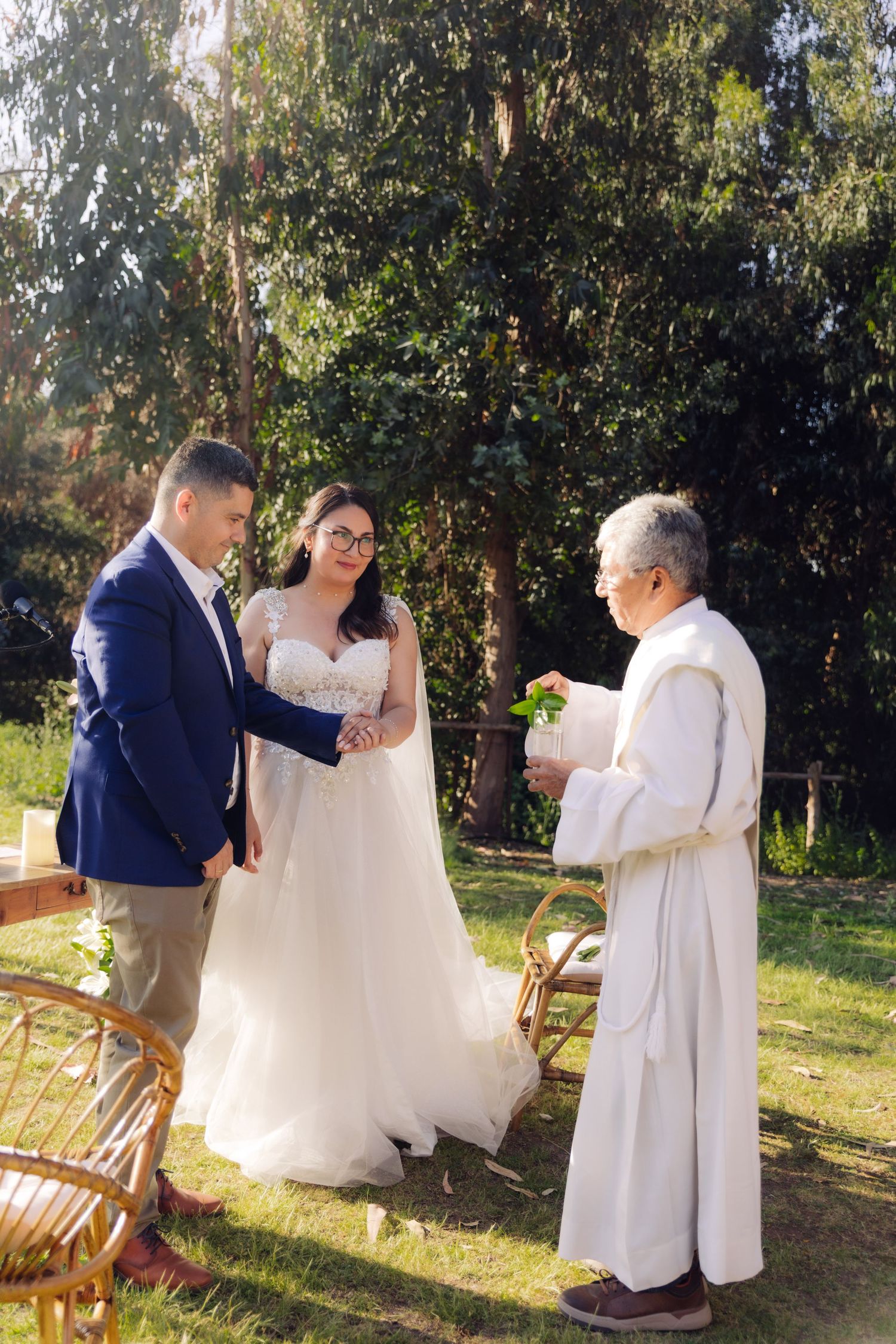 Bride and groom hold hands during outdoor wedding ceremony with officiant in white robes among green trees.