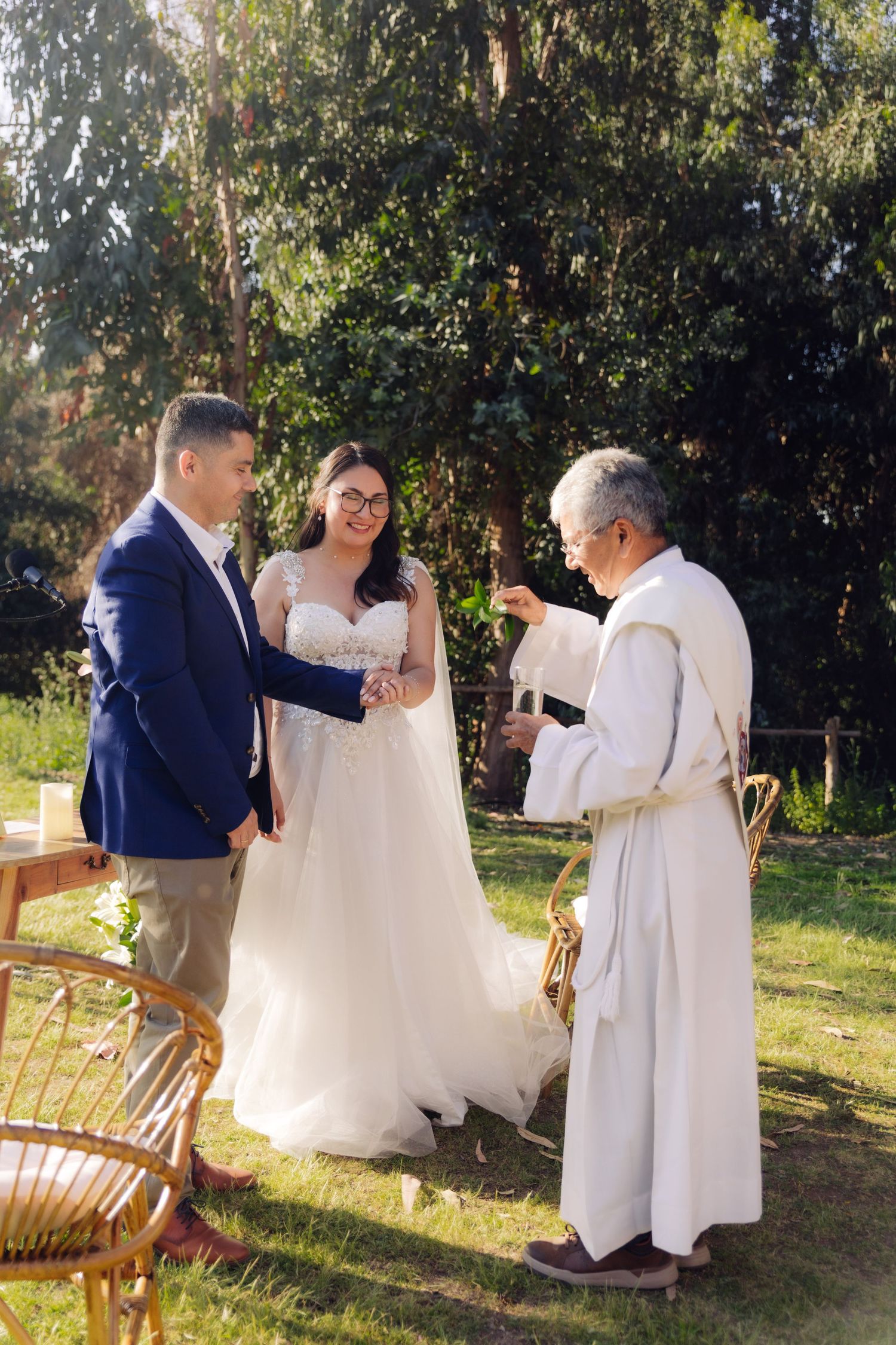 Smiling bride and groom exchange vows with robed officiant during sunny outdoor garden wedding ceremony.