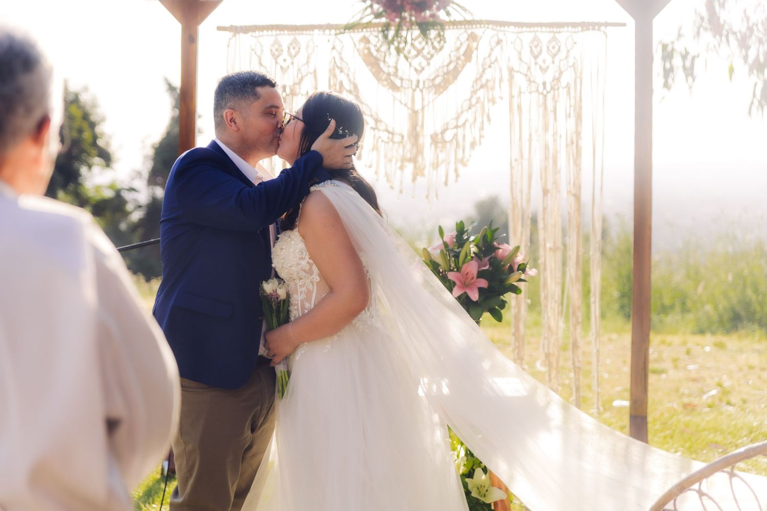 Newlywed couple shares first kiss under boho macrame arch with bride holding pink floral bouquet.