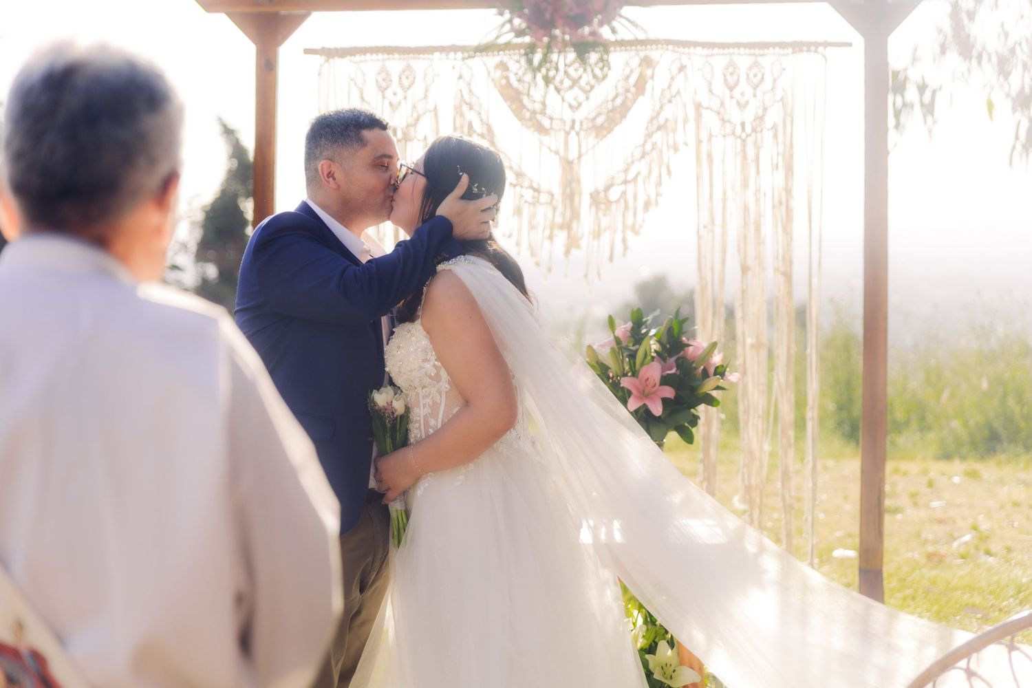 Bride and groom share romantic first kiss under decorative macrame wedding arch in bright sunlight.