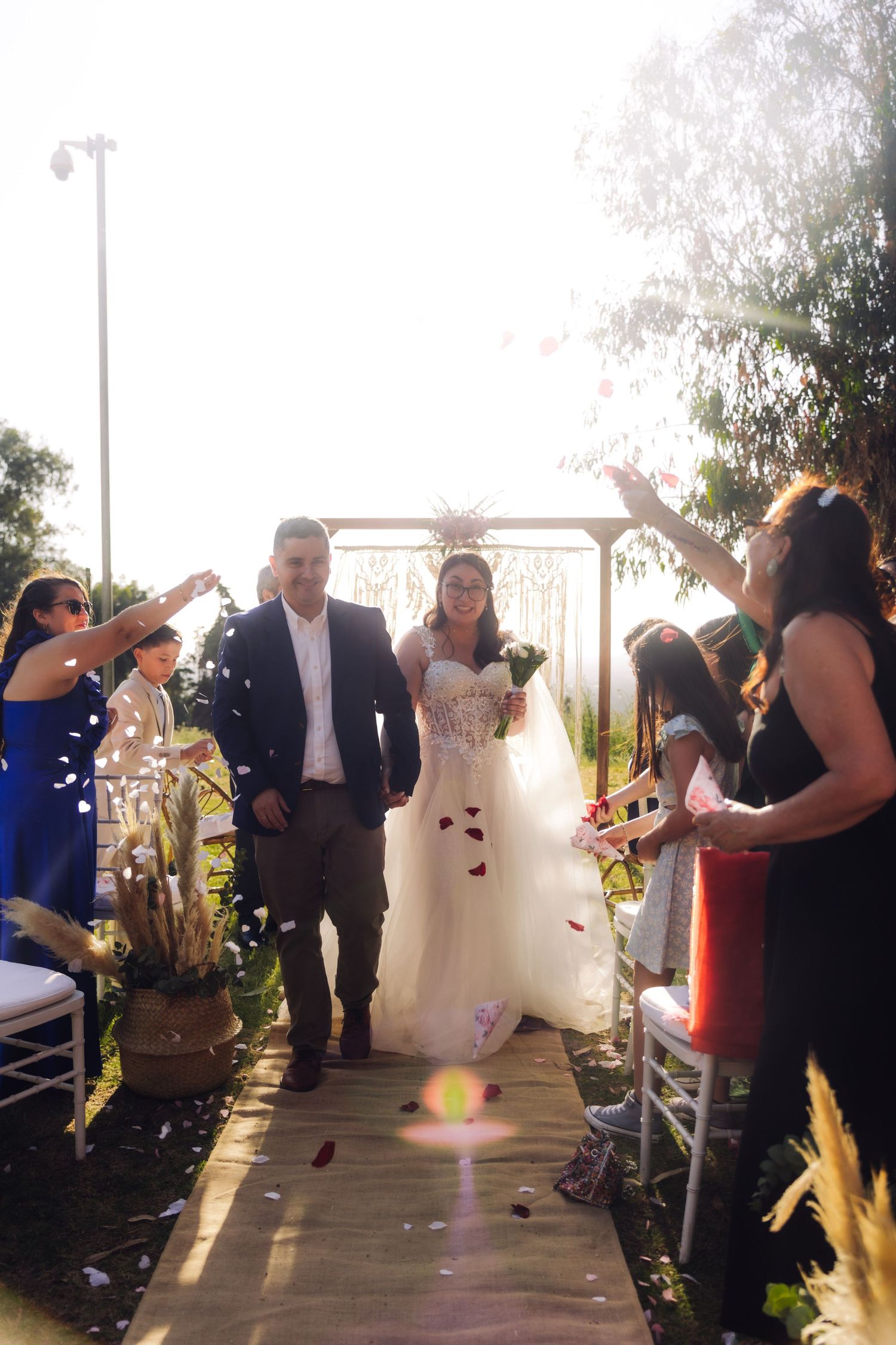 Joyful newlyweds walk down aisle as guests toss confetti and rose petals at outdoor wedding ceremony.