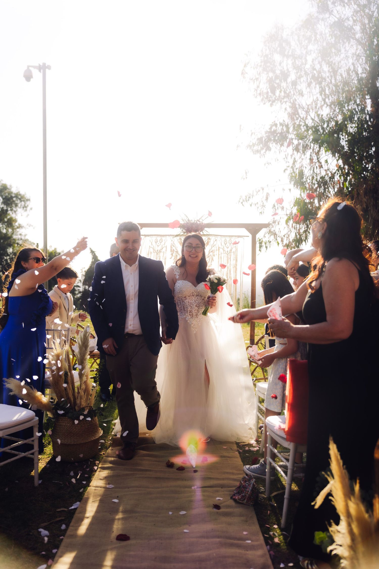 Bride and groom walk through confetti shower as guests celebrate along sunlit outdoor wedding aisle.