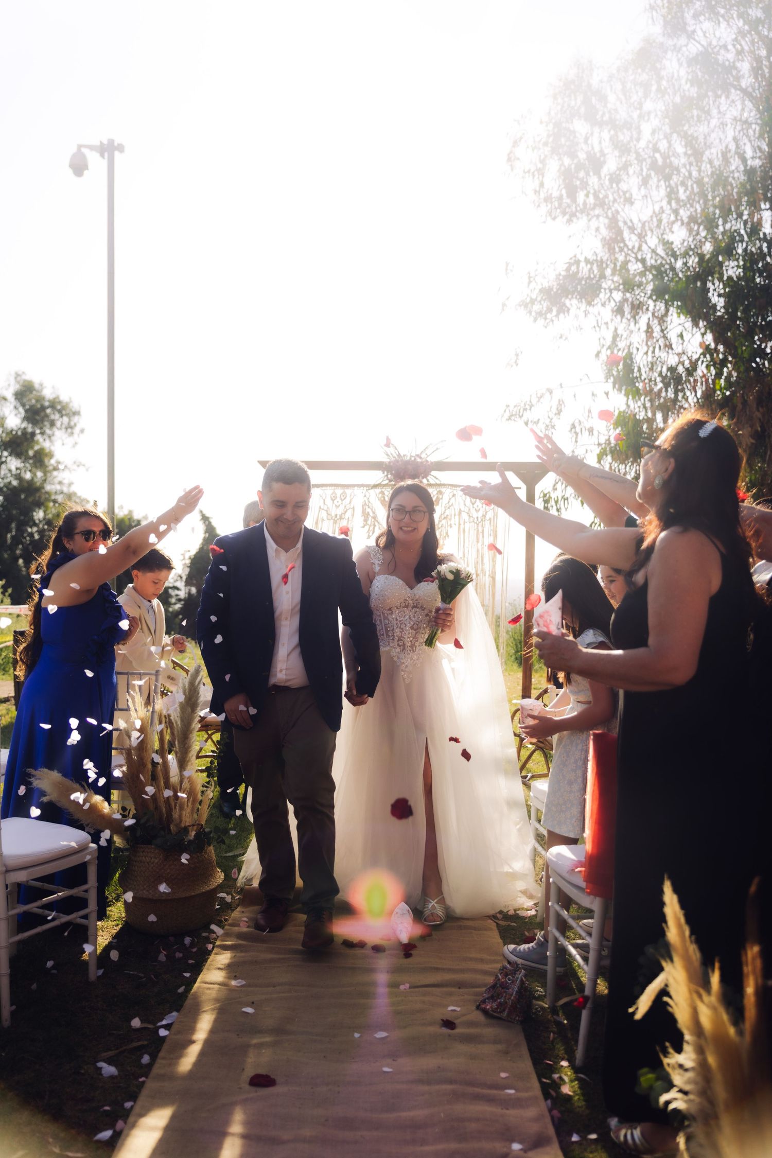 Newlywed couple walks down sunlit outdoor aisle while smiling guests toss confetti in celebration.