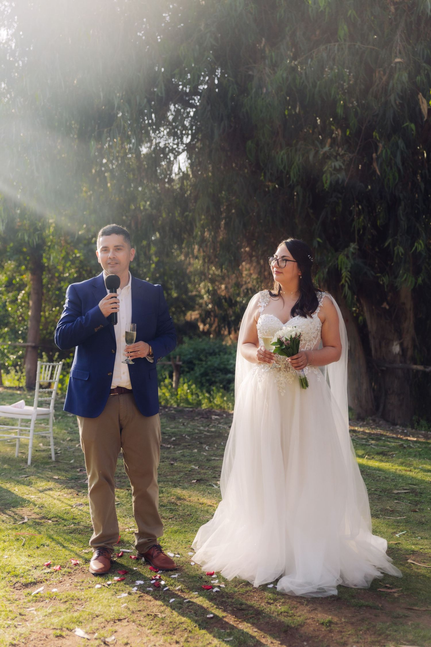 Groom speaks into microphone holding champagne glass beside bride with bouquet at outdoor wedding.
