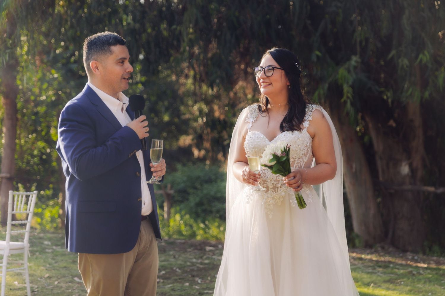 Groom toasts with champagne glass while laughing bride holds bouquet during outdoor wedding reception.