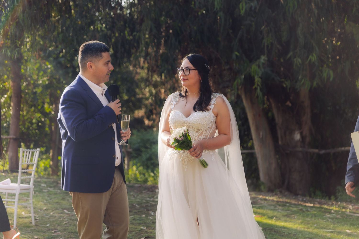 Bride smiles joyfully as groom speaks with champagne glass at sunny outdoor wedding celebration.