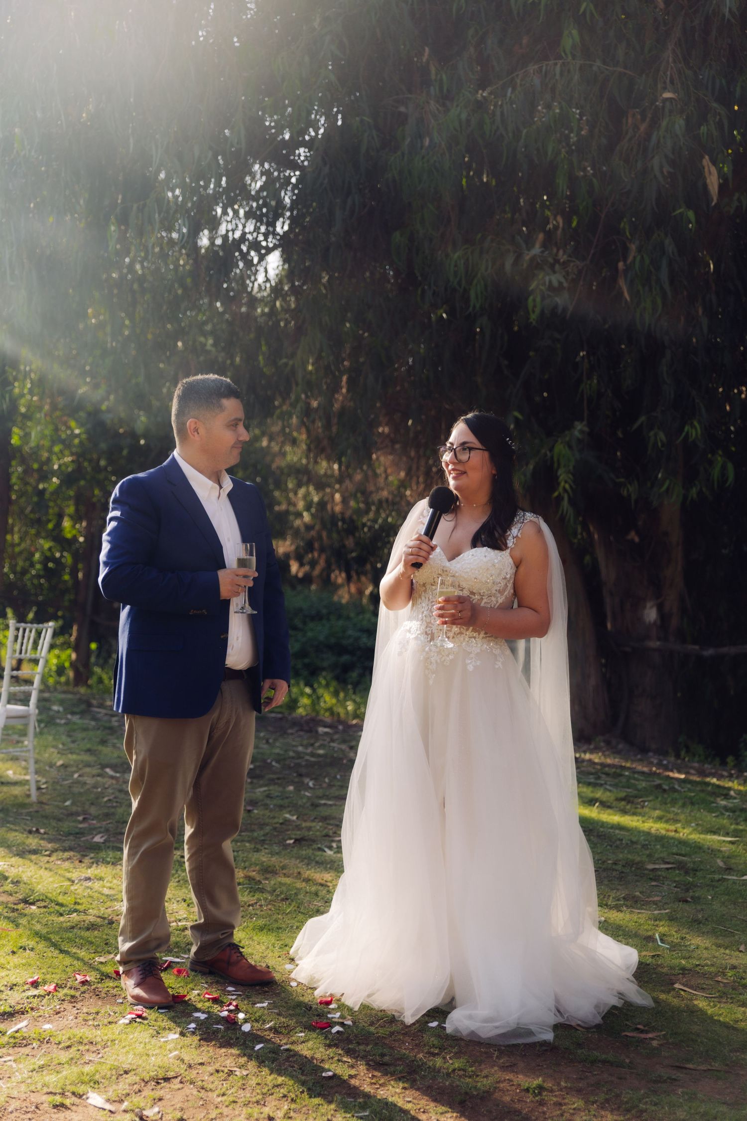 Bride and groom stand outdoors in sunlit garden, she in white gown holding mic, he in blue blazer with champagne.