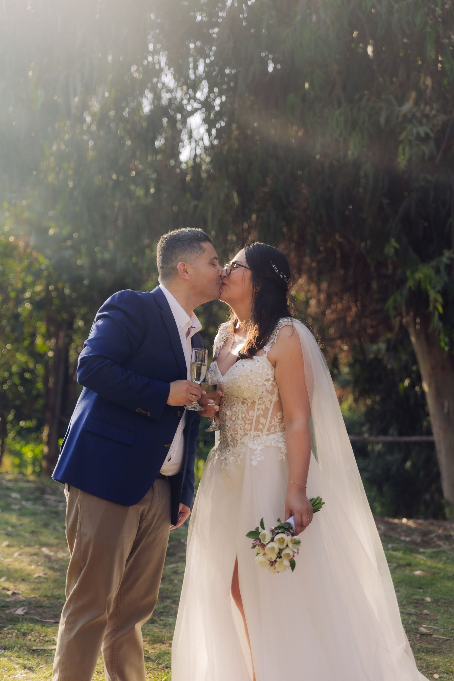 Newlyweds share a kiss outdoors in golden sunlight, bride in white gown with veil holding bouquet and champagne.