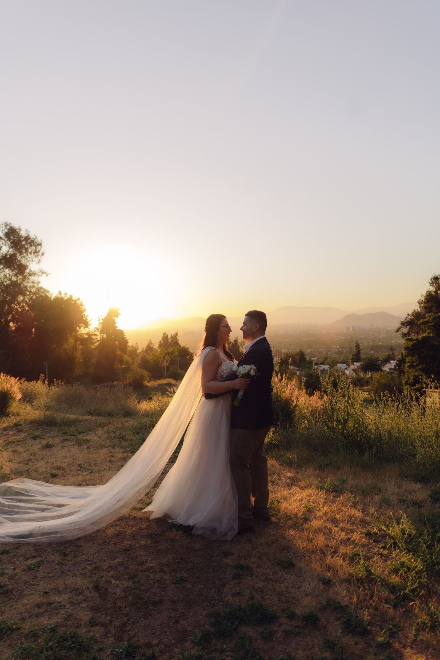 Bride and groom embrace at sunset on hilltop, her long veil flowing in wind with panoramic landscape behind them.