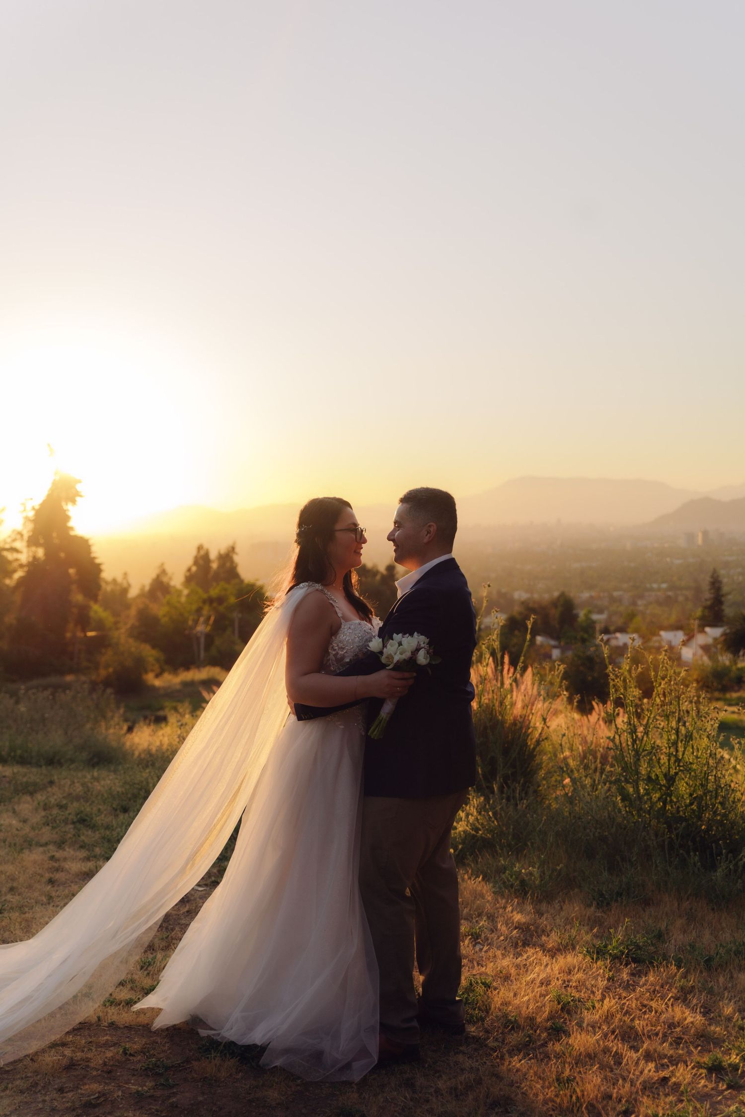 Wedding couple gazes lovingly at each other during golden sunset on scenic hilltop, bride holding white bouquet.
