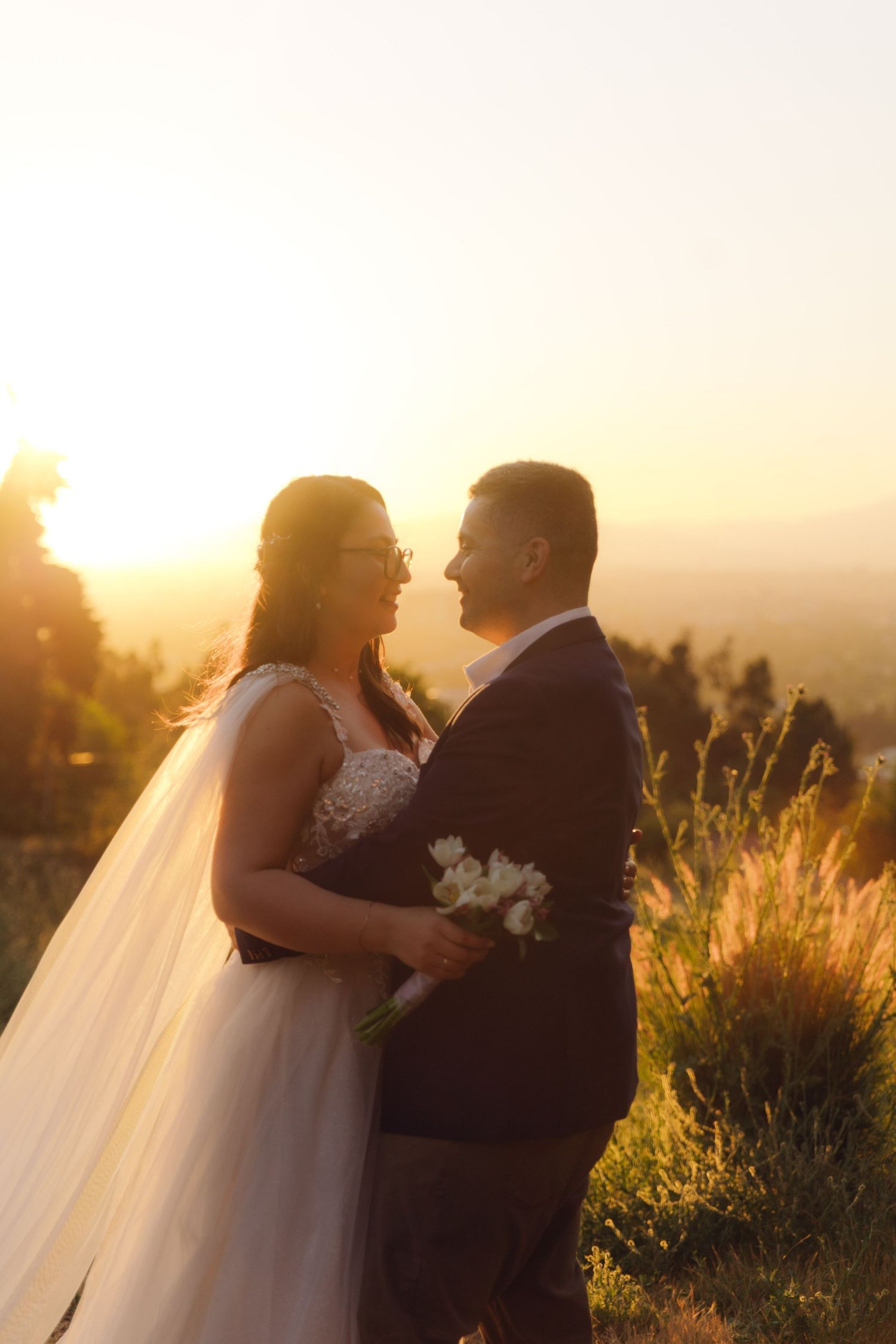 Bride and groom share intimate moment at golden sunset, she holding white flowers, mountains visible in background.